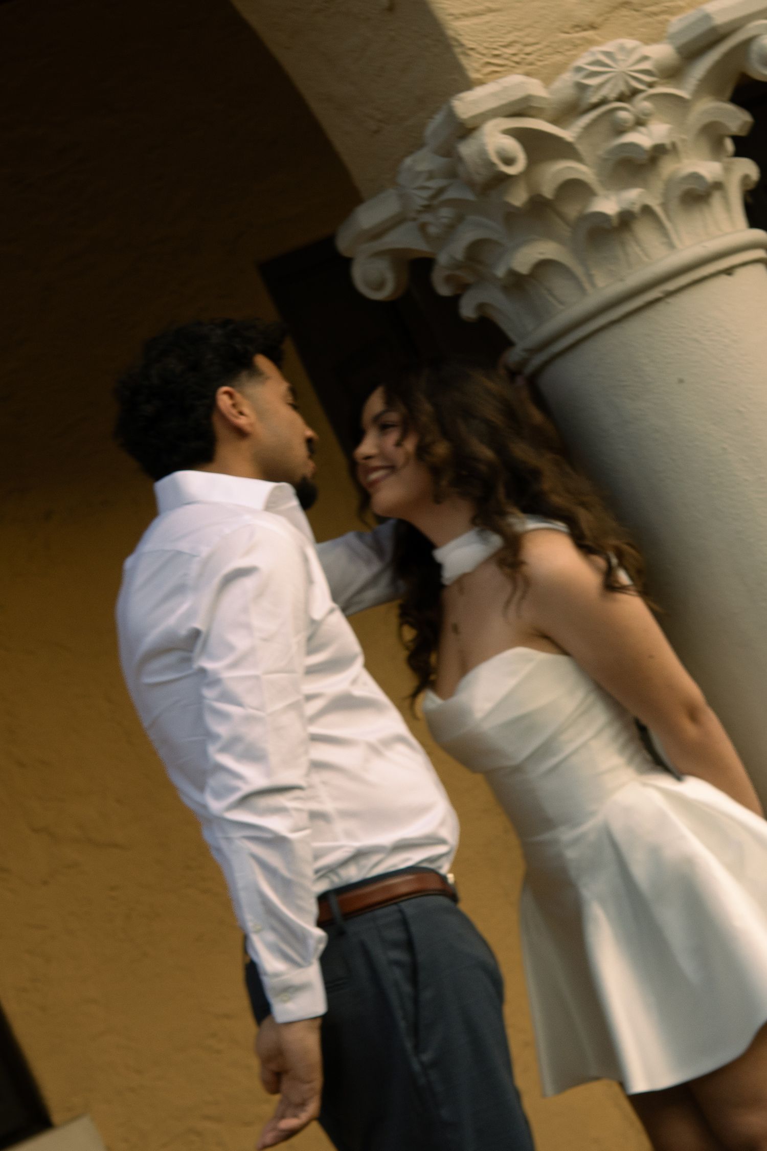 Couple in white outfits sharing a romantic moment beside an ornate stone column.