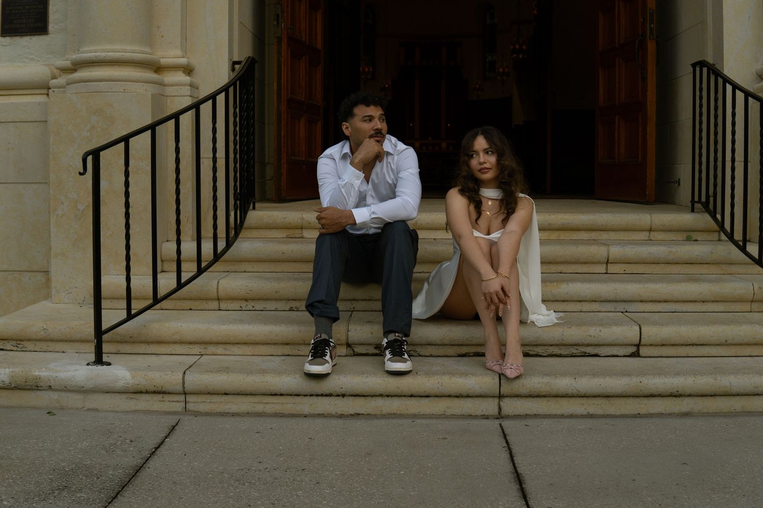 Couple sitting apart on church steps, woman in white mini dress, man in white shirt and jeans.