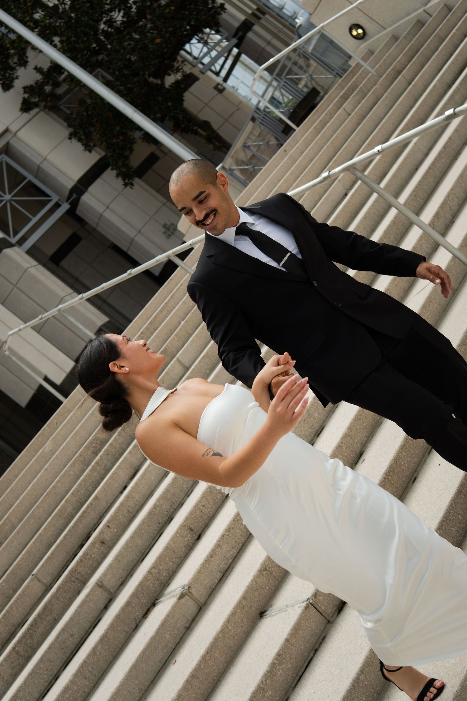 Smiling groom in black suit holds bride's hands on outdoor steps during their wedding photo session.