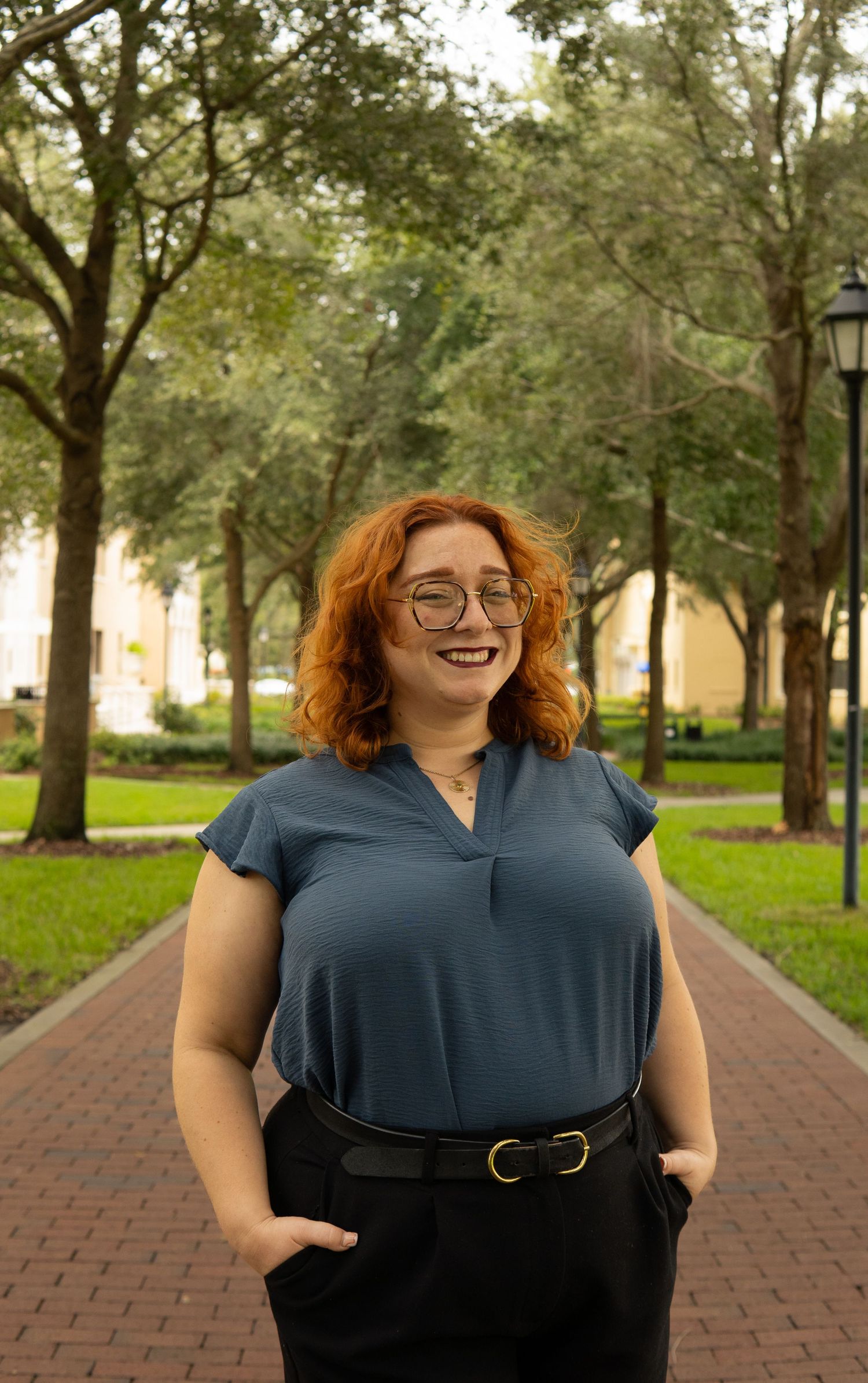 Smiling woman with red hair and glasses wearing a blue top and black pants on a brick pathway.