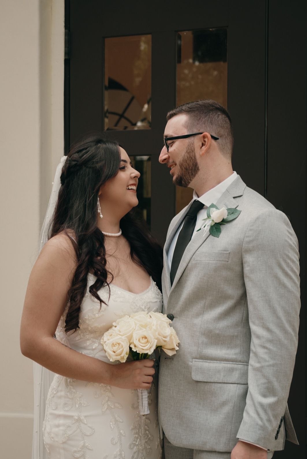 Bride holding white rose bouquet gazes at groom in gray suit outside dark doors.