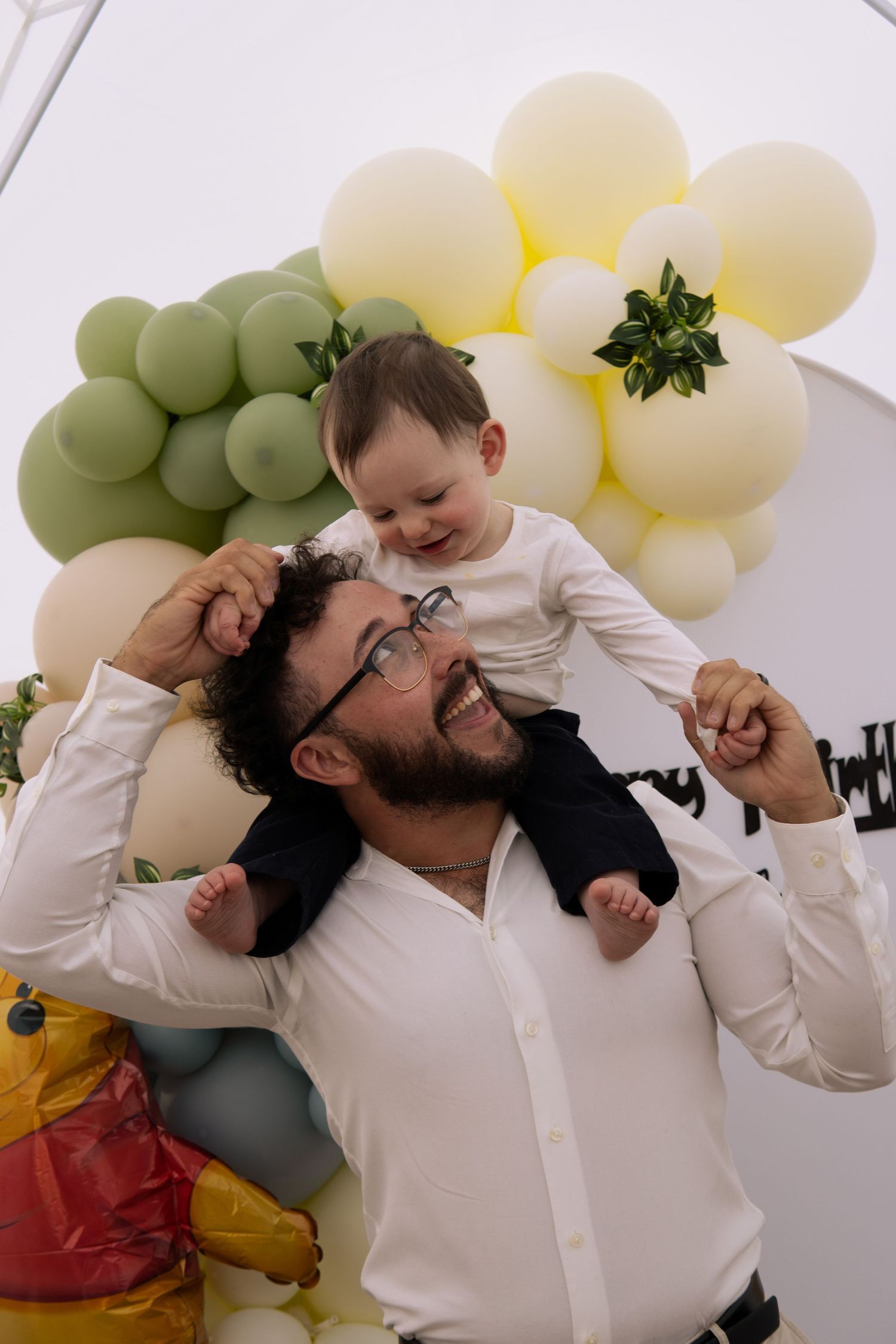 Smiling father holds laughing baby on shoulders in front of colorful balloon backdrop.