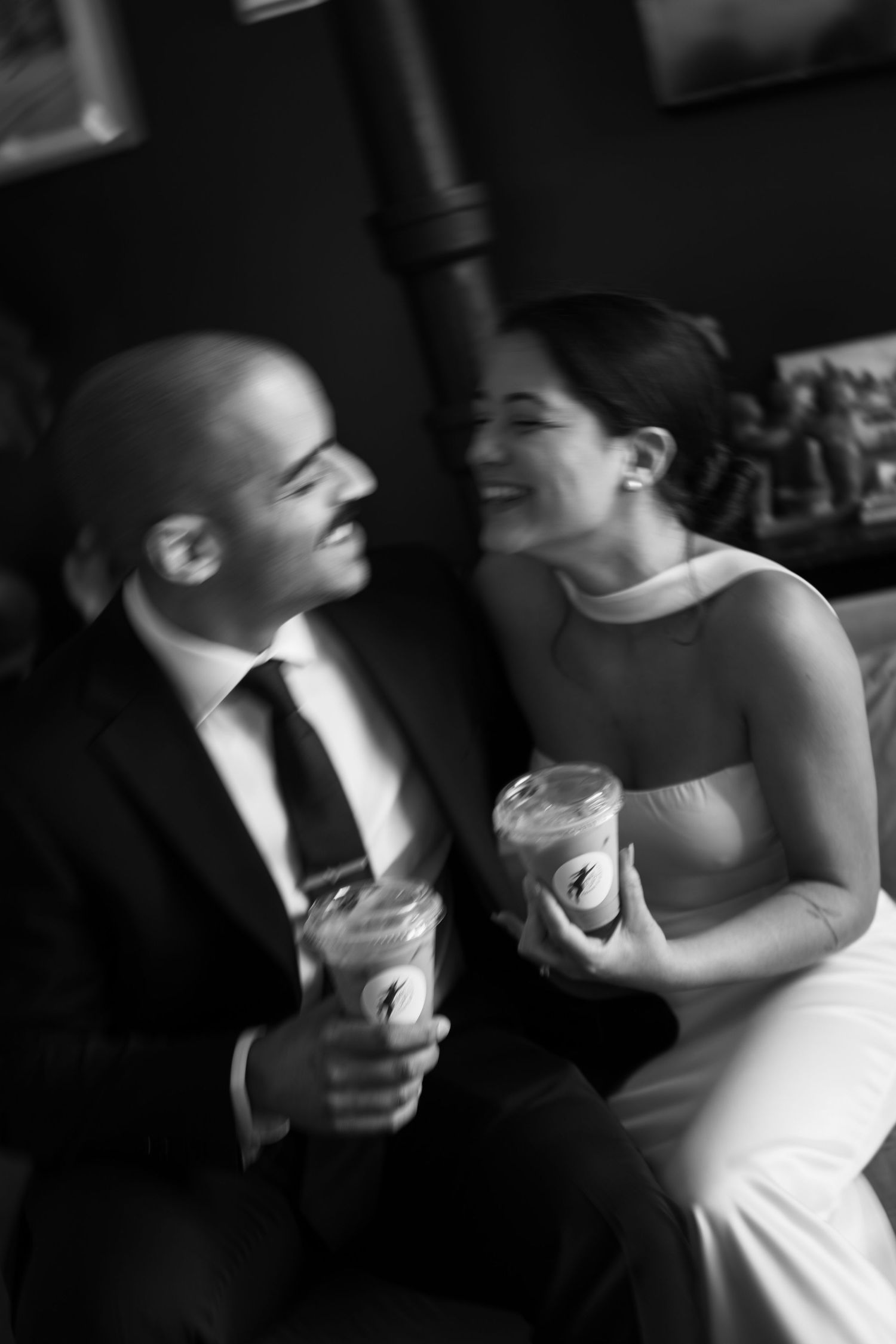 Black and white photo of bride and groom laughing together while holding drinks at wedding reception.