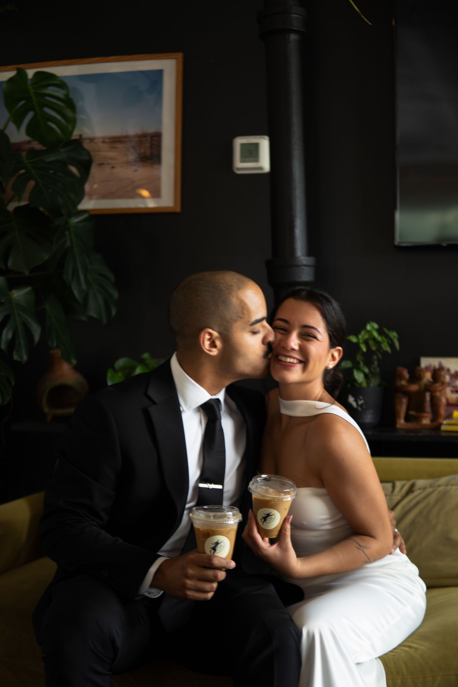 Newlywed couple in wedding attire sharing iced coffees with a kiss on a green sofa.