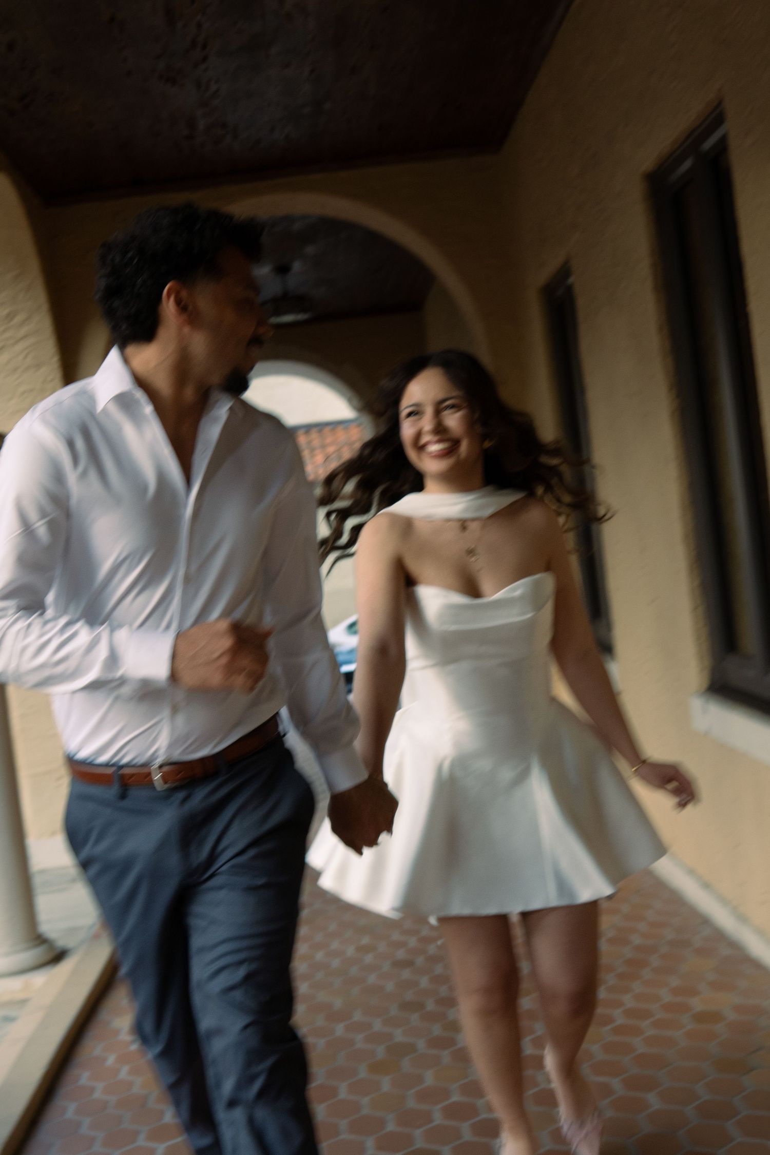 Bride and groom running hand in hand through an arched corridor, laughing joyfully on their wedding day.