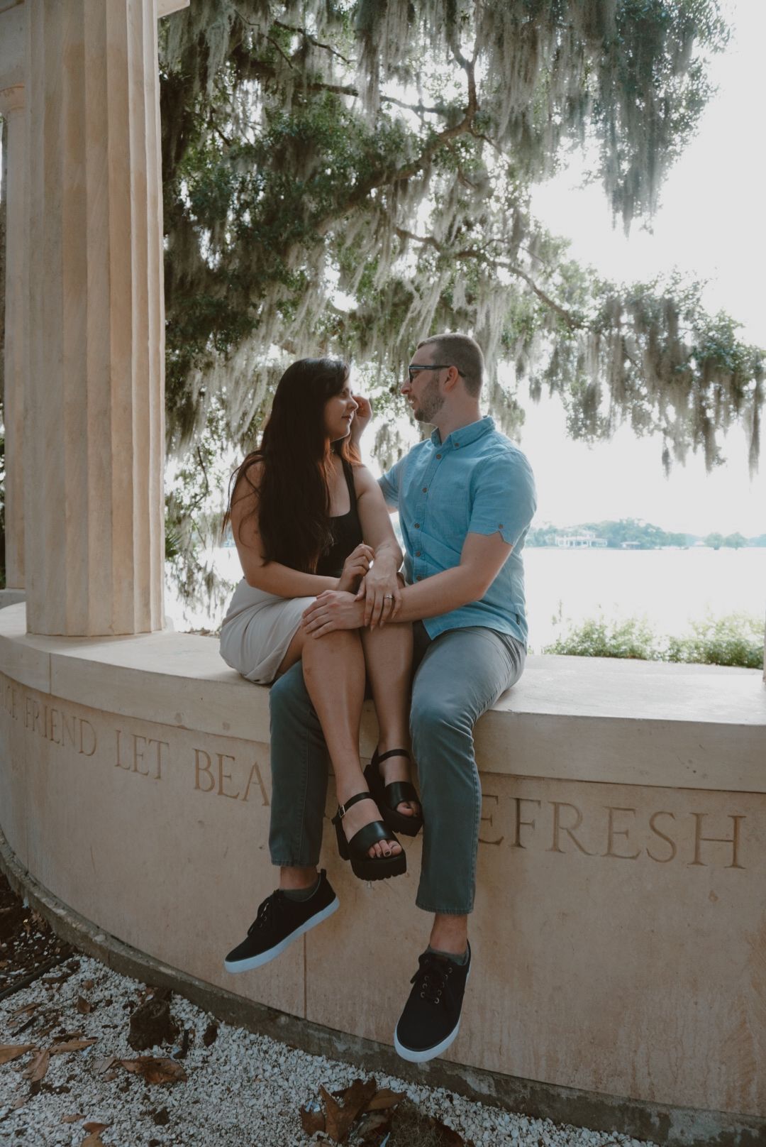 Engaged couple sits together on a marble bench with lake views and Spanish moss trees.