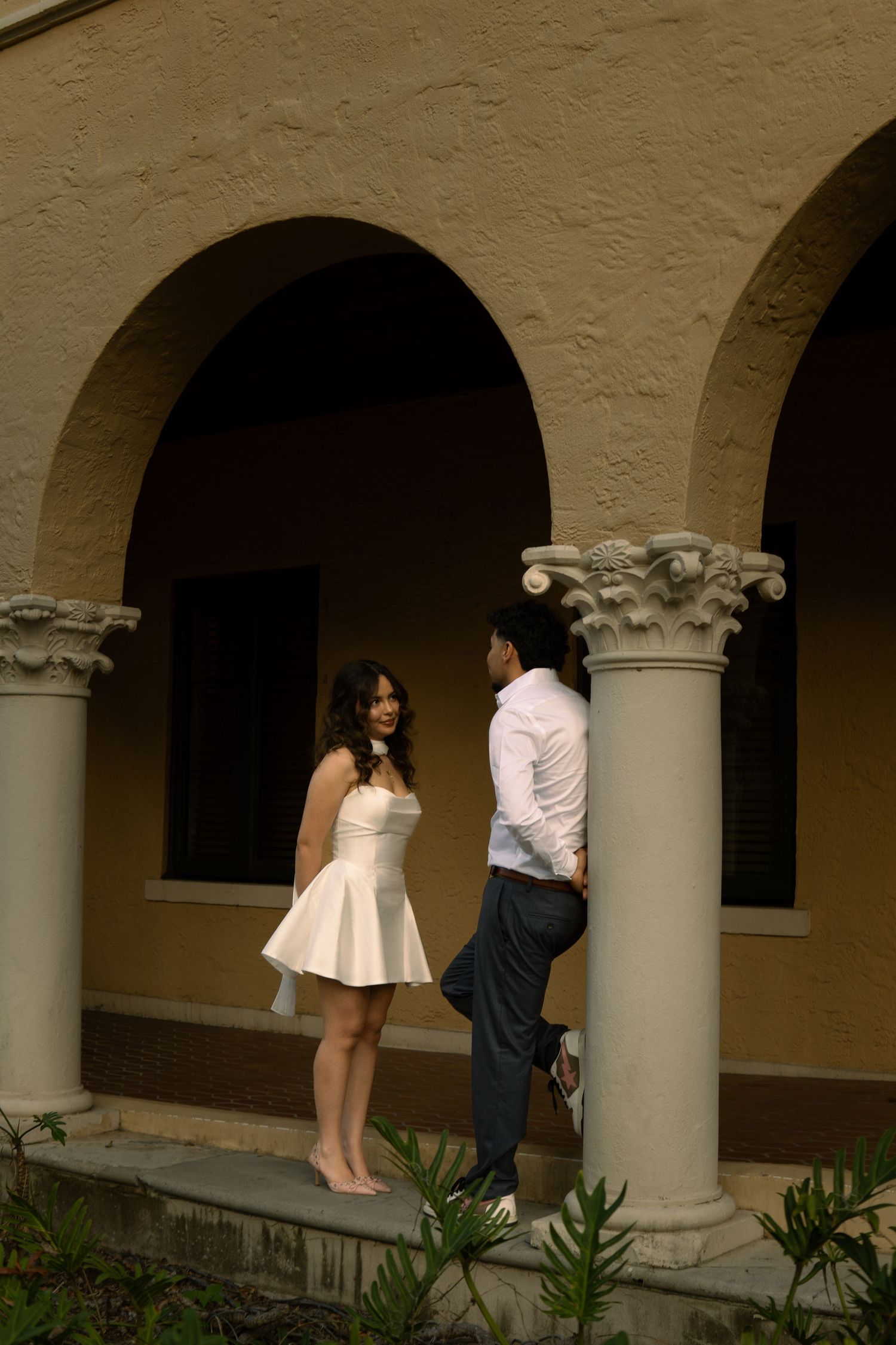 Woman in white strapless mini dress facing man in white shirt under decorative arched colonnade.
