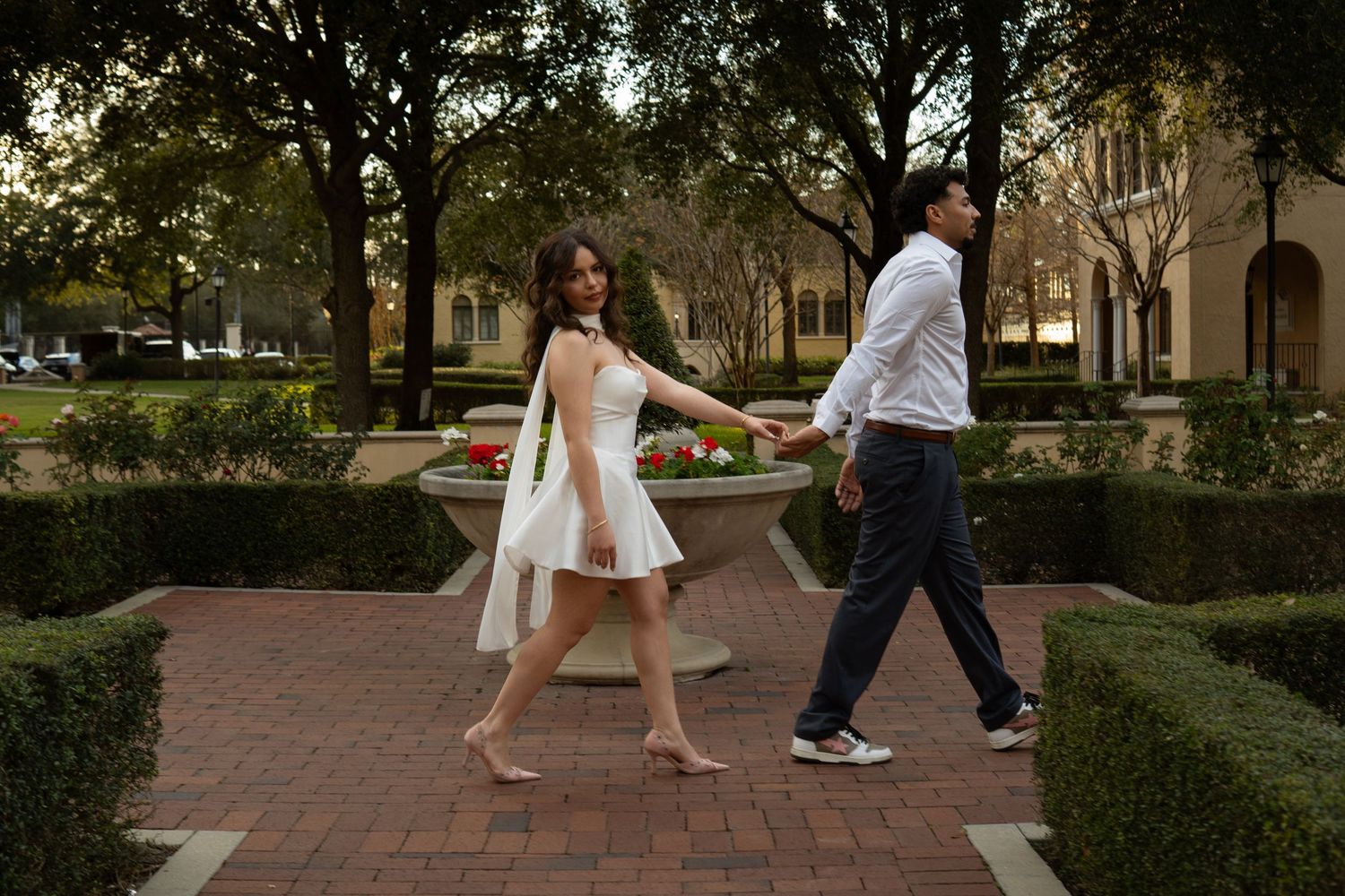 Couple walking hand in hand through a brick garden path, woman in white dress leading the way.