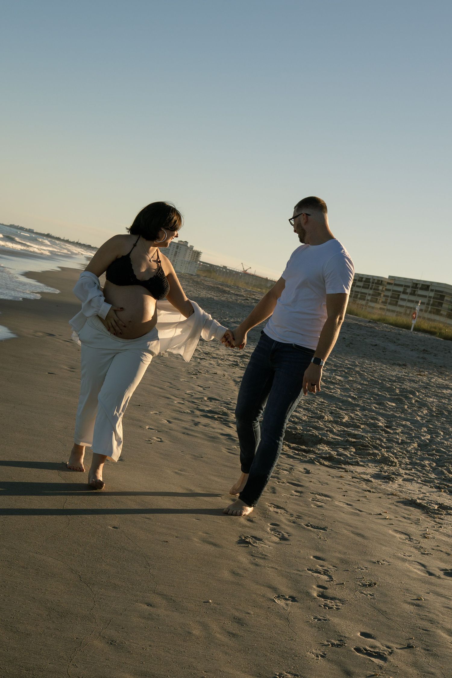 A couple walks along a sandy beach at sunset, the pregnant woman cradling her baby bump.