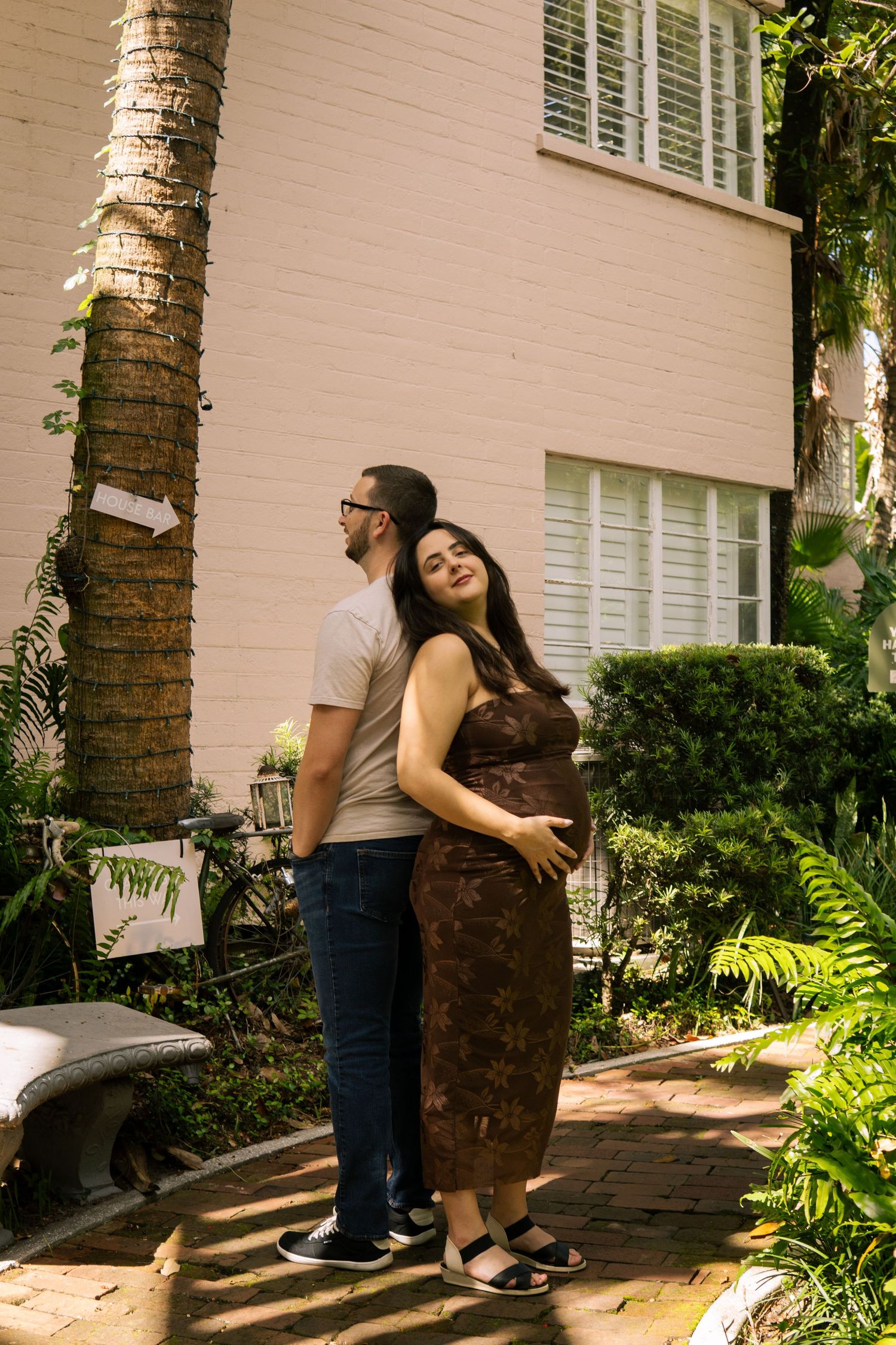 Pregnant couple posing in front of a pink building surrounded by lush tropical greenery.