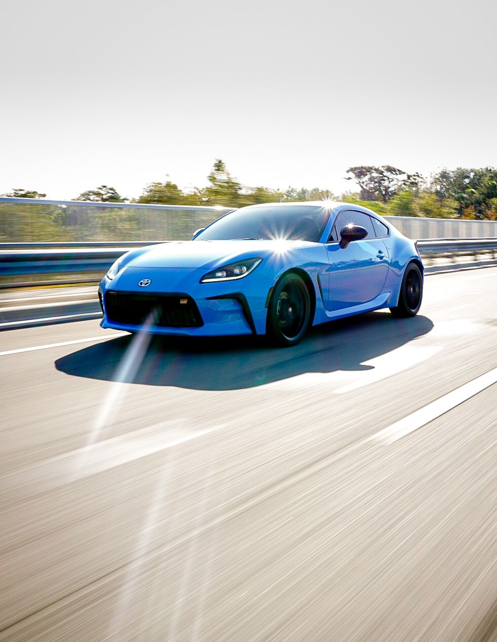 A vibrant blue Toyota GR86 sports car speeds along a highway with motion blur background.
