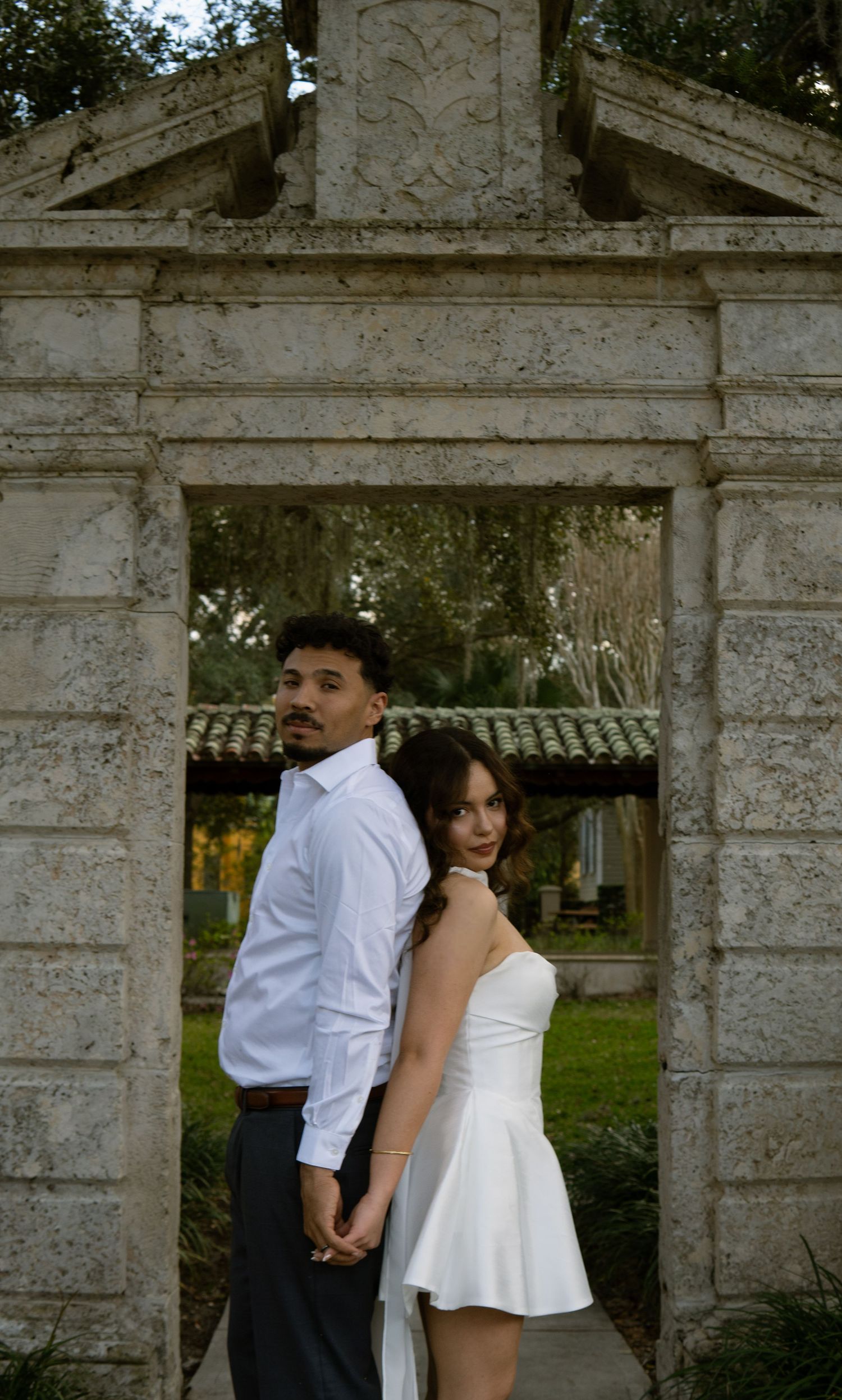 Couple holding hands back-to-back framed by ornate stone garden gate archway outdoors.