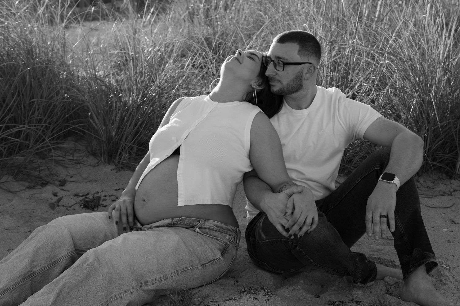 Black and white photo of a couple sitting on sand dunes, woman leaning on tattooed man in a tender moment.