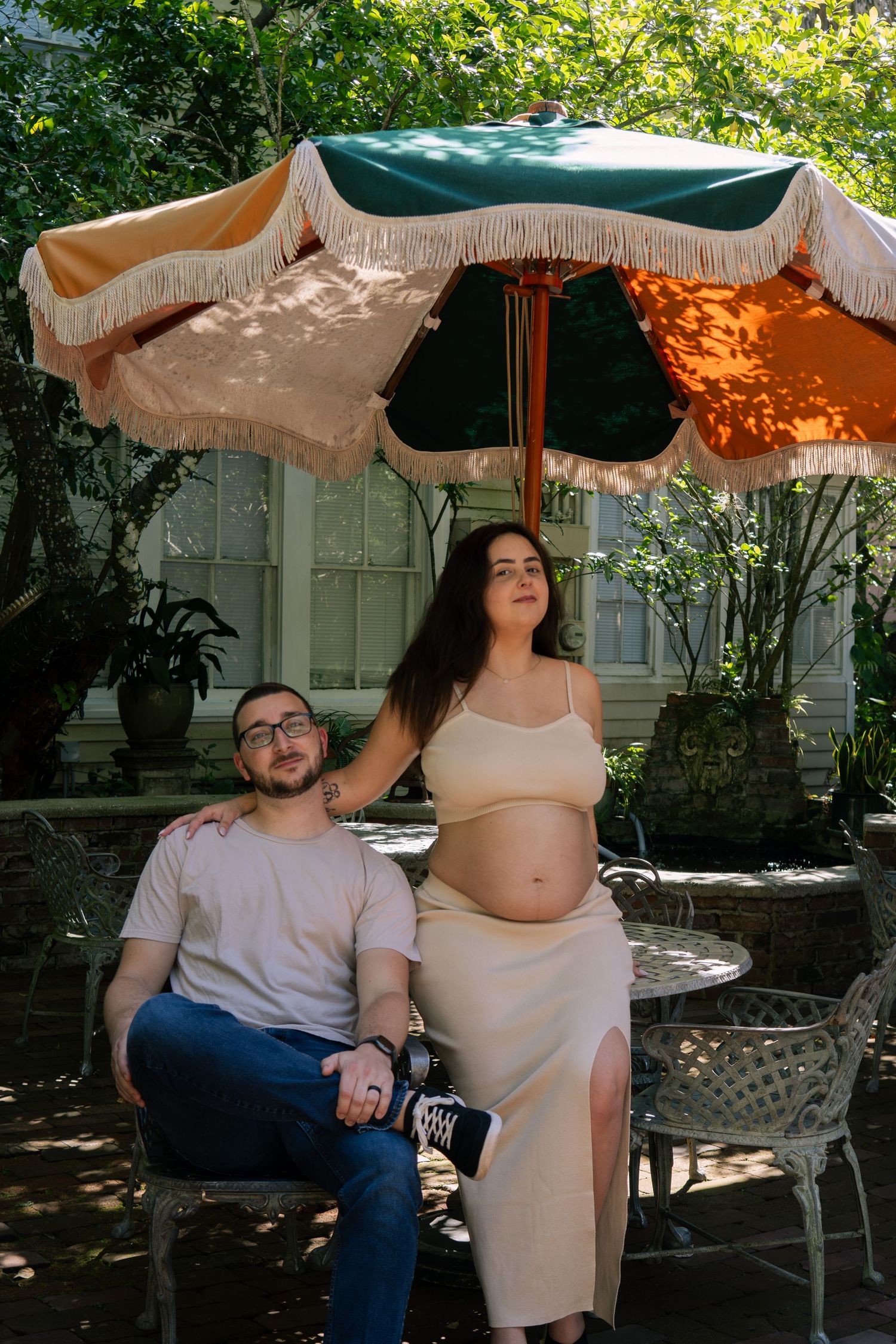 Expecting couple relaxes under fringed patio umbrella at outdoor garden table in sunlight.