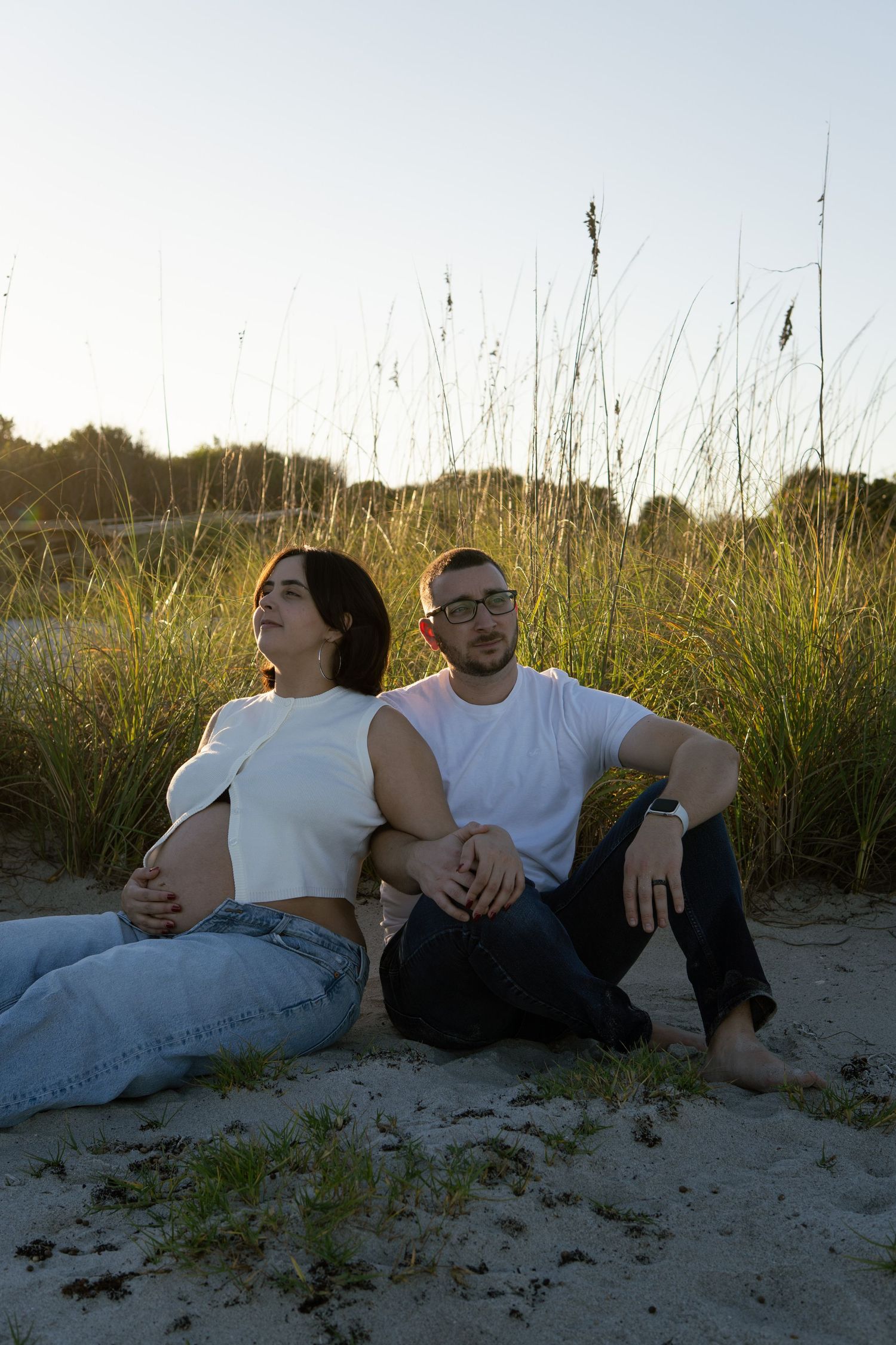 A couple sits back to back on a sandy beach surrounded by tall grass at sunset.