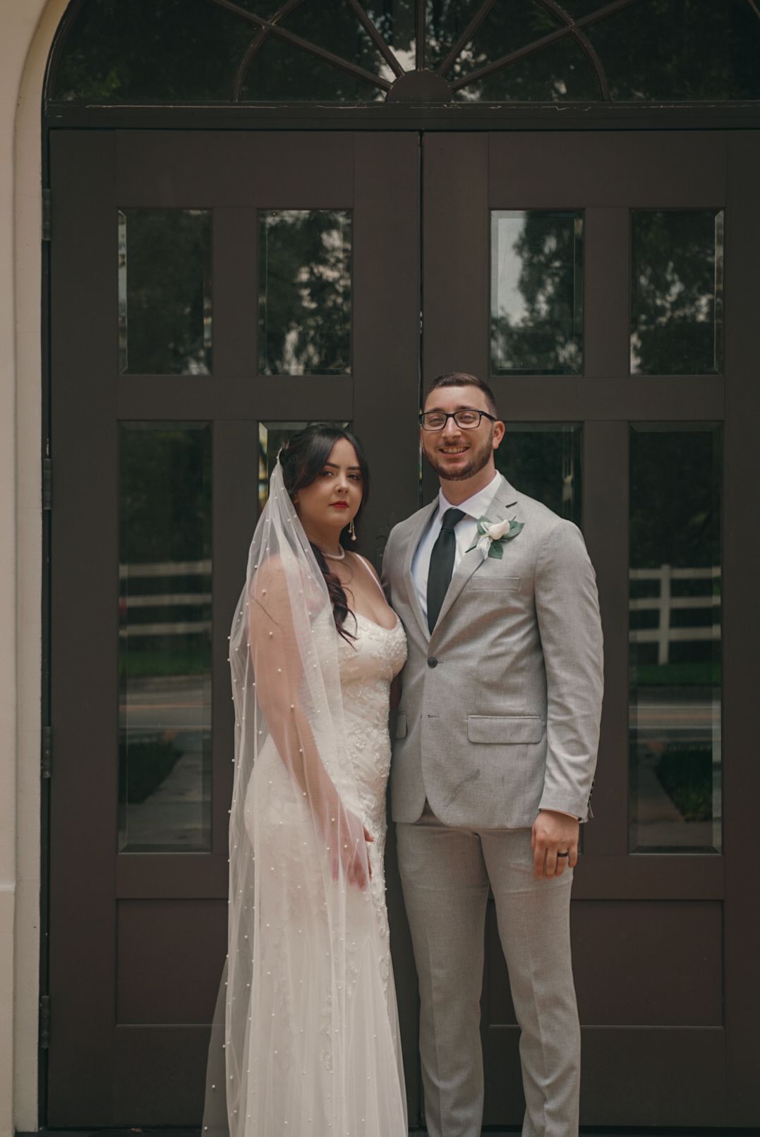 Bride in white lace gown with pearl veil and groom in gray suit posing before ornate dark wooden doors.