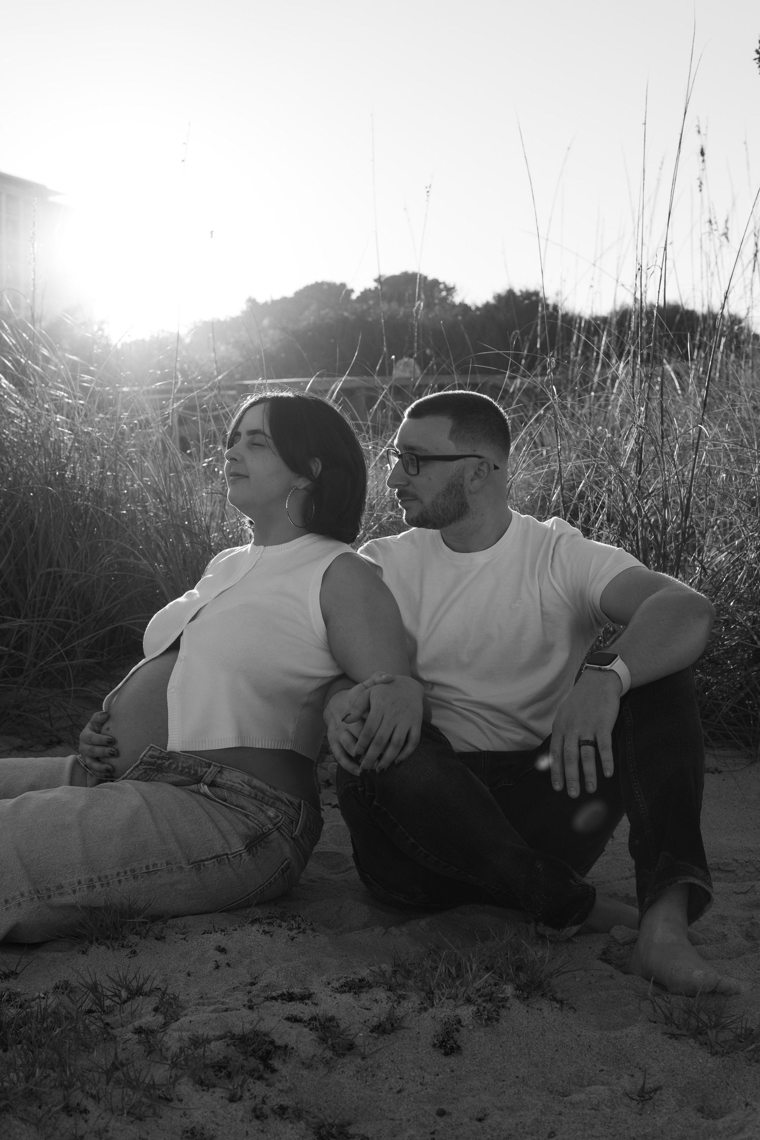 Couple sits back-to-back on sandy ground in black and white sunset portrait.
