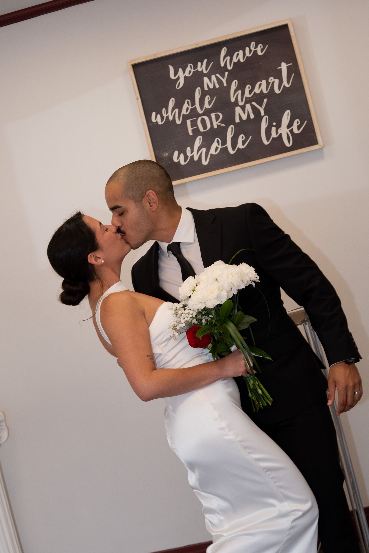Newlywed couple sharing a romantic kiss, bride holding white bouquet, with a love quote sign in the background.