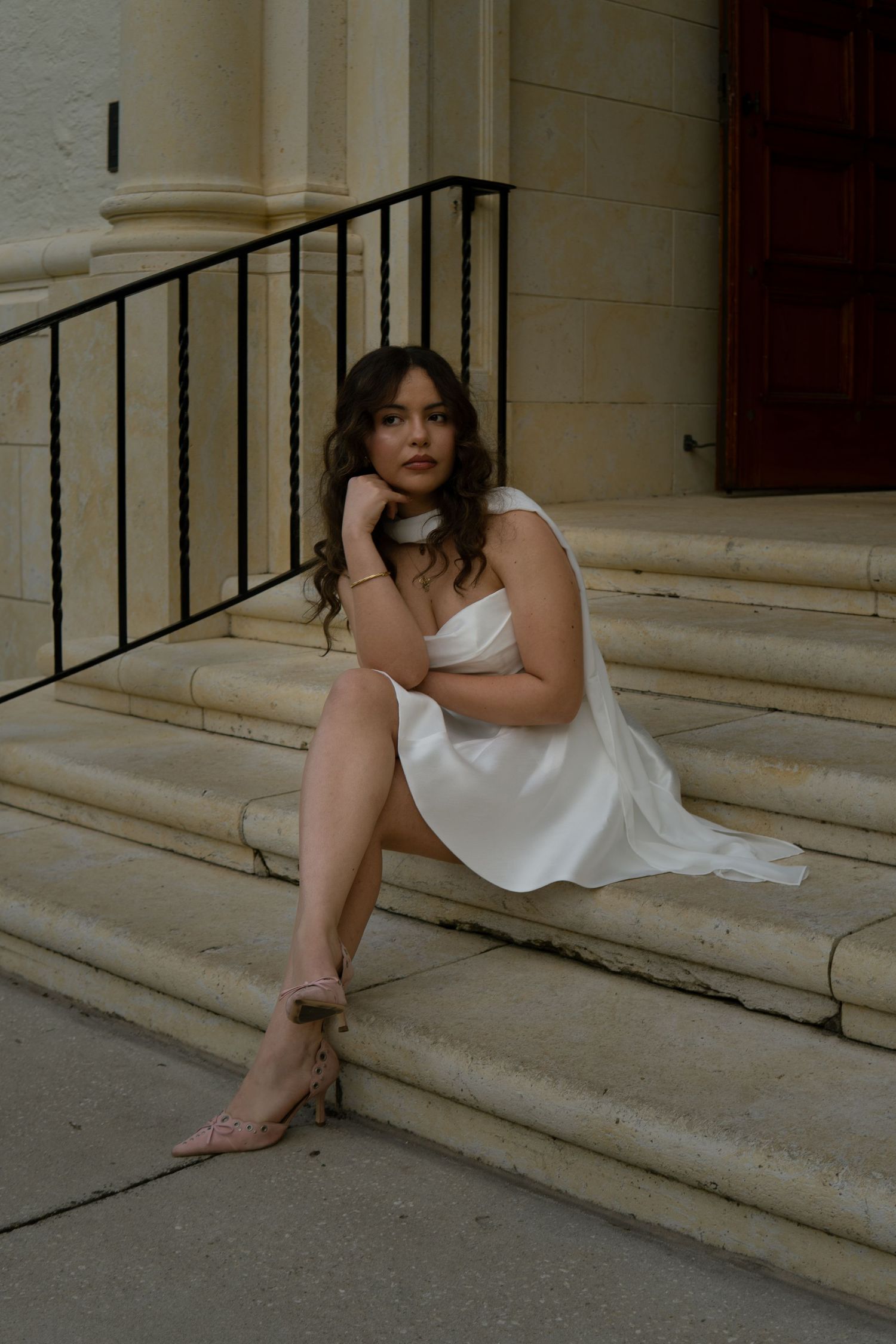 Bride in a white satin mini dress seated on stone steps wearing nude heels in a contemplative pose.