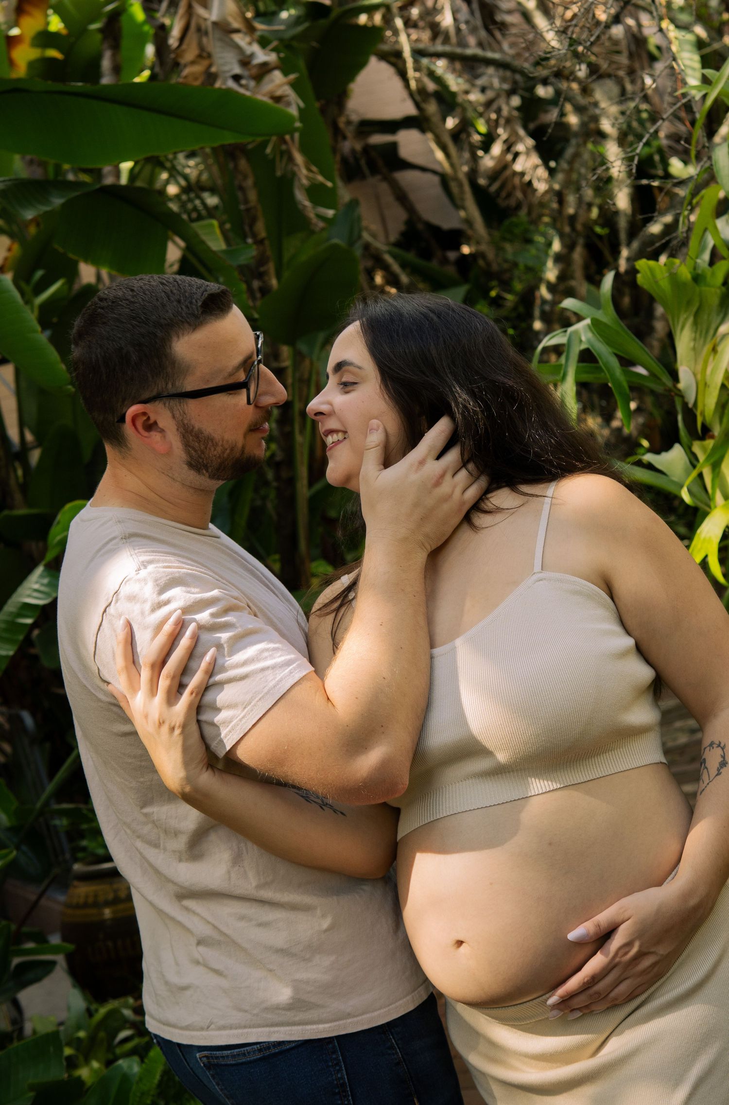 Smiling expecting couple embraces outdoors surrounded by lush tropical green foliage.