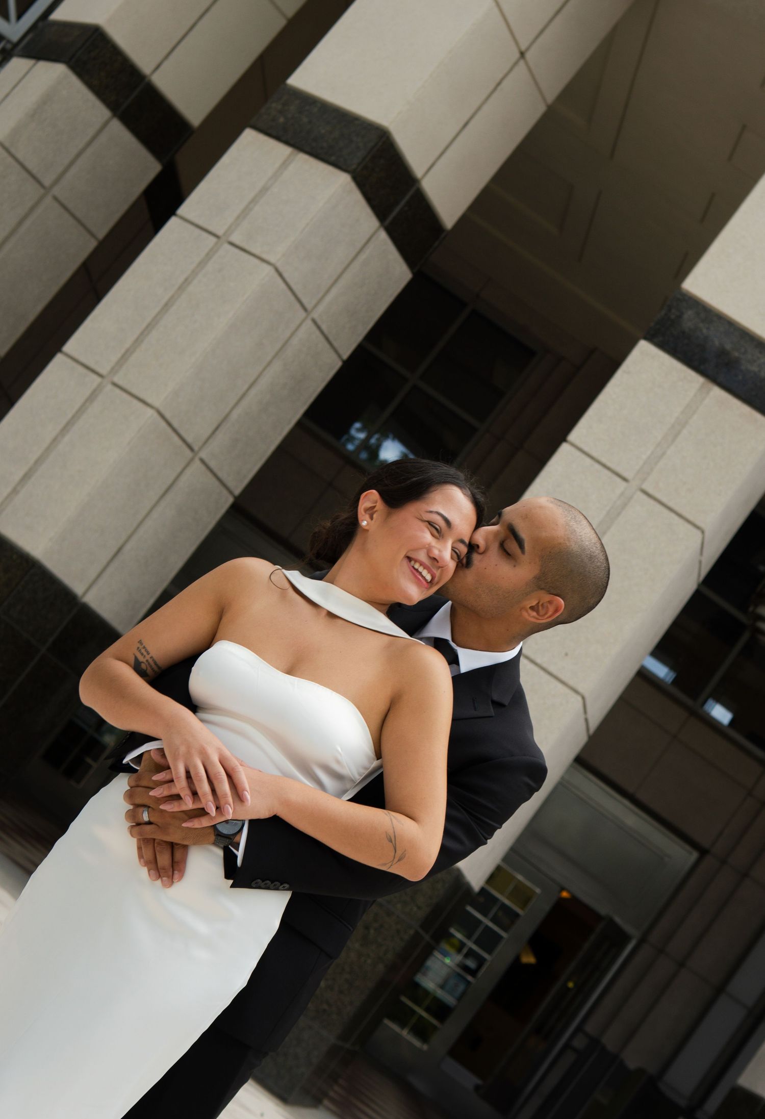 Groom in black suit kissing smiling bride in white strapless gown outdoors near modern building columns.