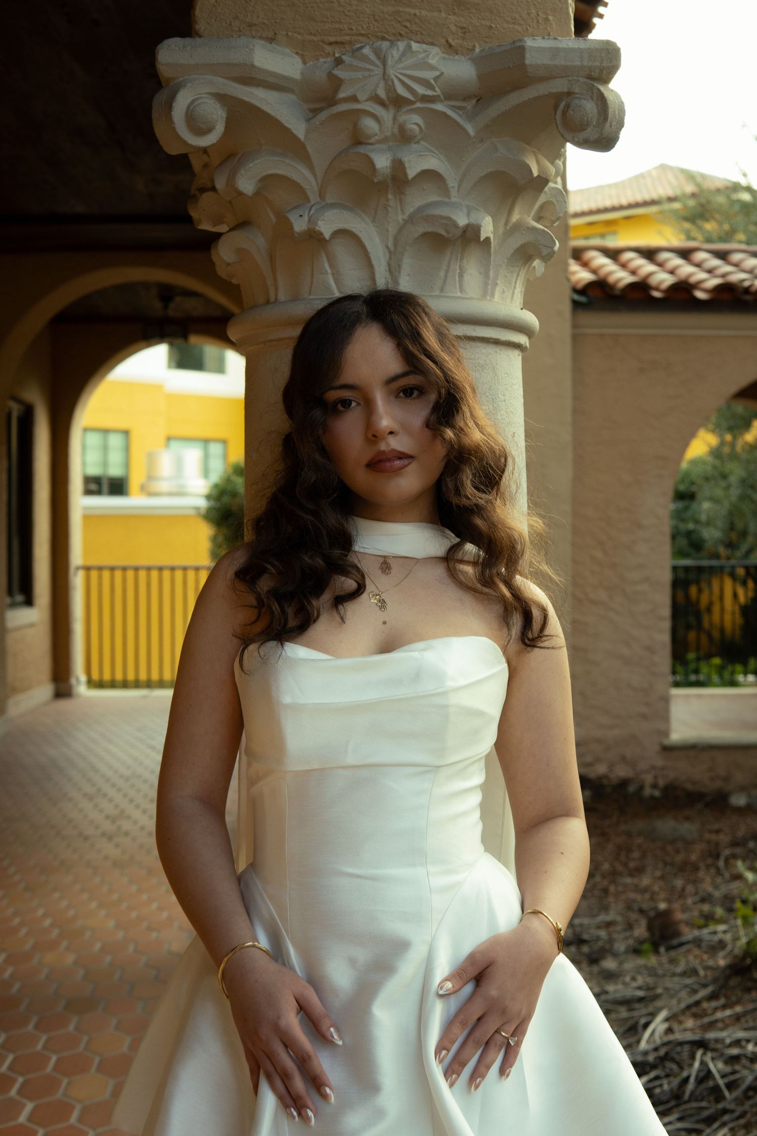 Elegant woman in white strapless gown posing beneath ornate Corinthian column in courtyard.