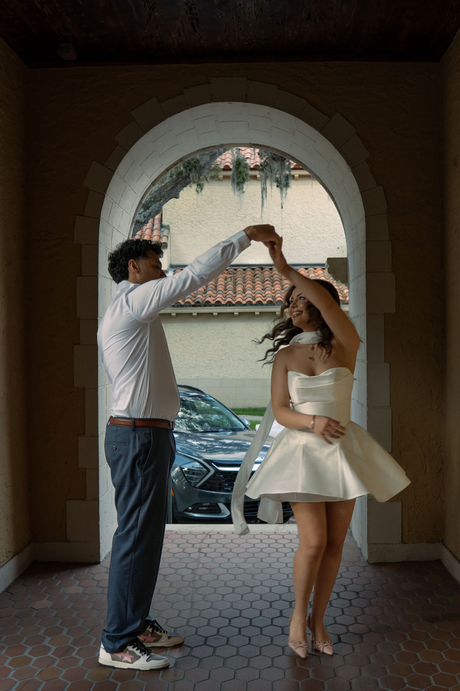 Couple dancing and spinning under stone archway with man twirling woman in white mini dress.