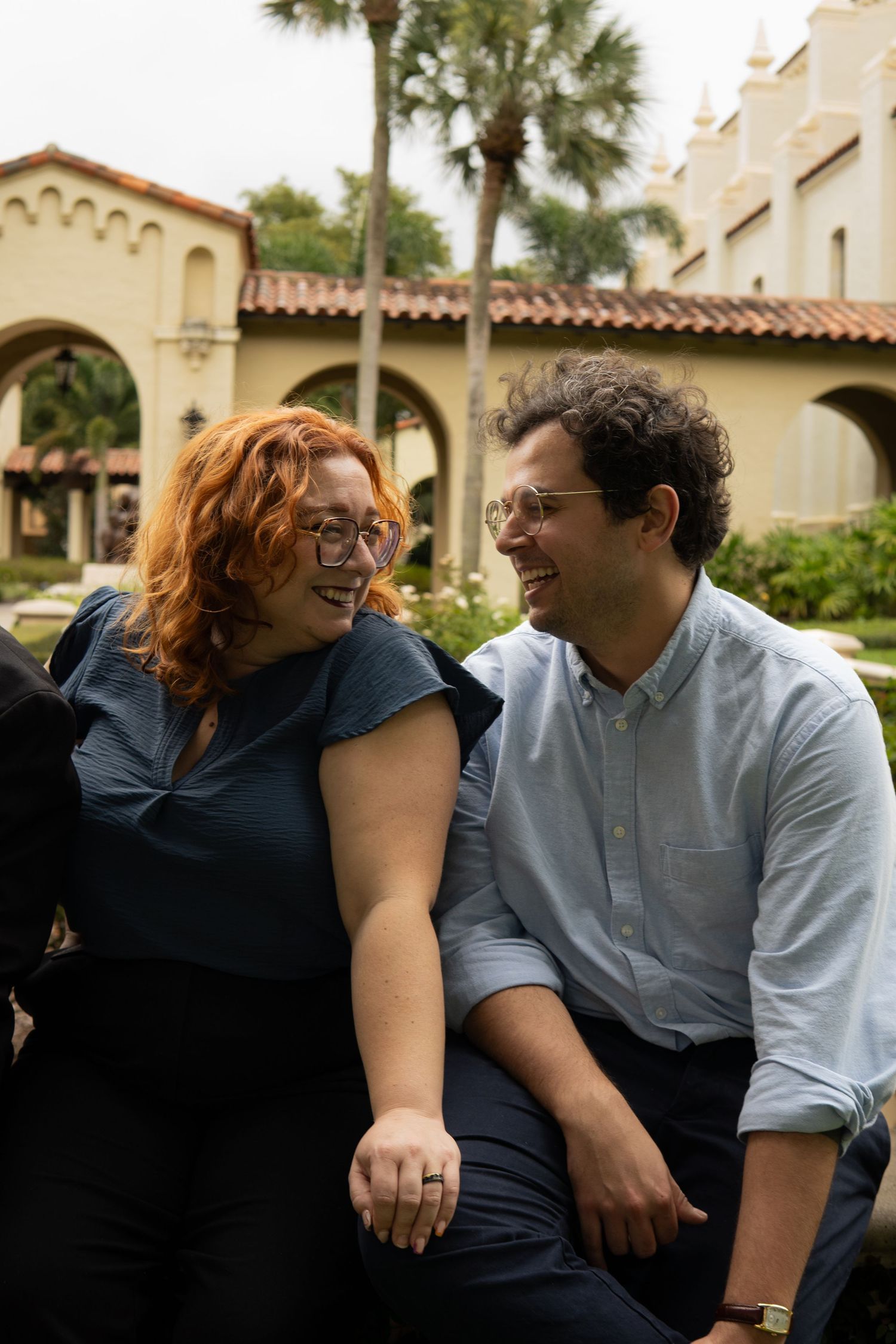 Red-haired woman and curly-haired man laughing together outdoors near arched building.
