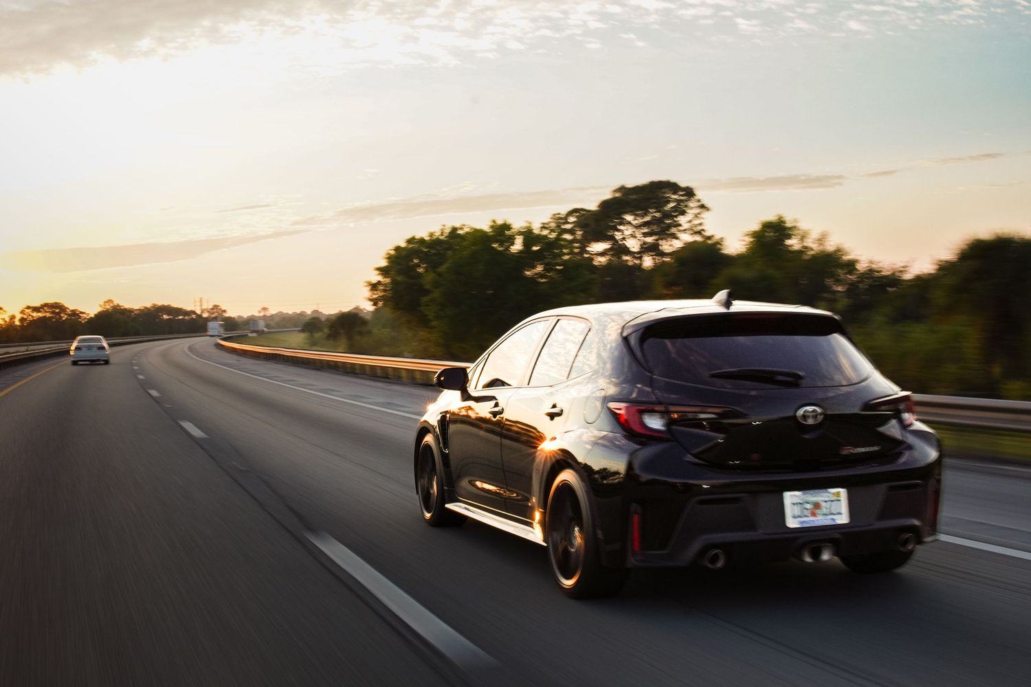 Black Toyota GR Corolla hatchback speeding on open highway during golden hour sunset.