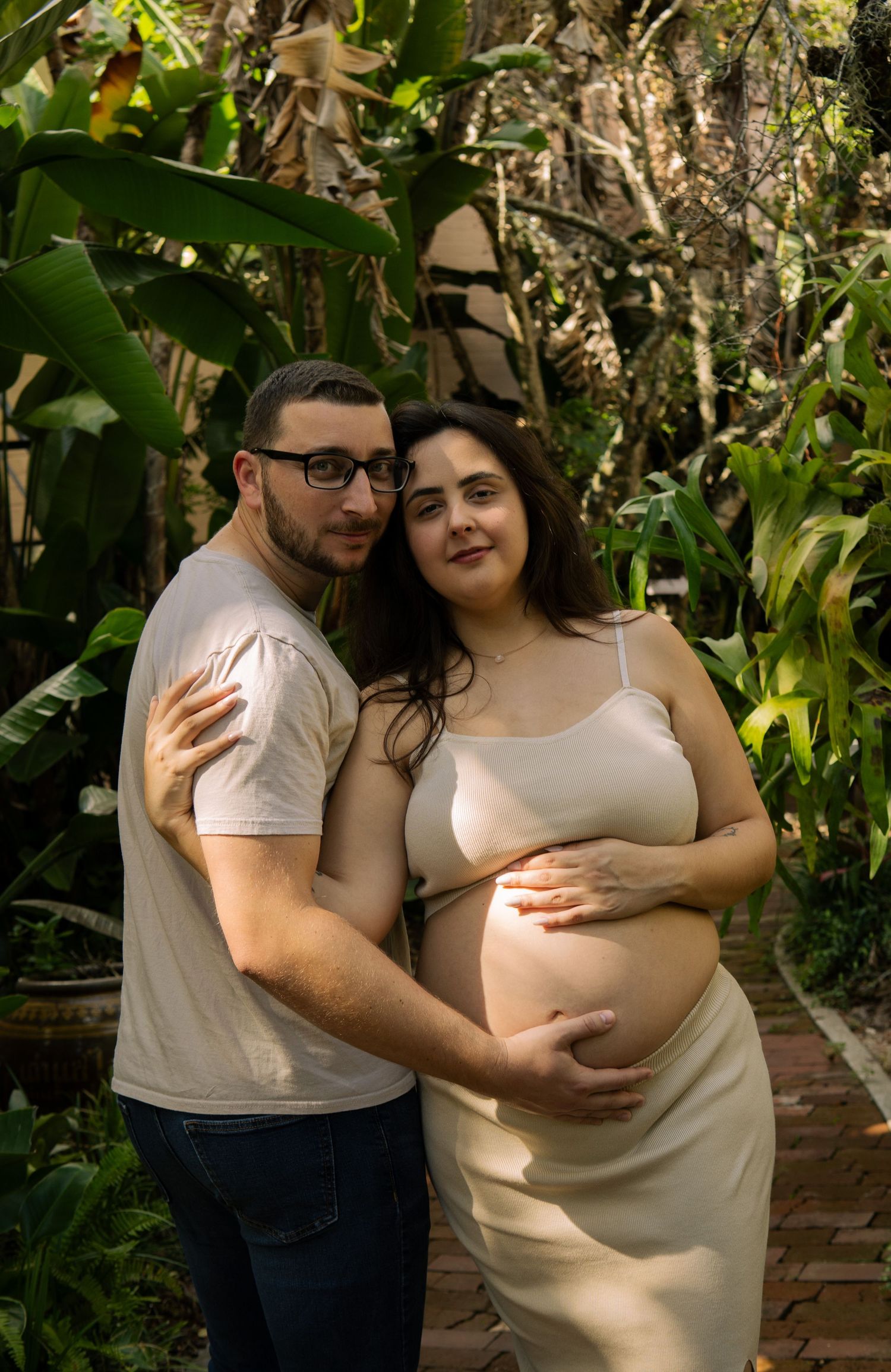 Expecting couple poses together in tropical garden, woman in beige maternity dress holding bump.