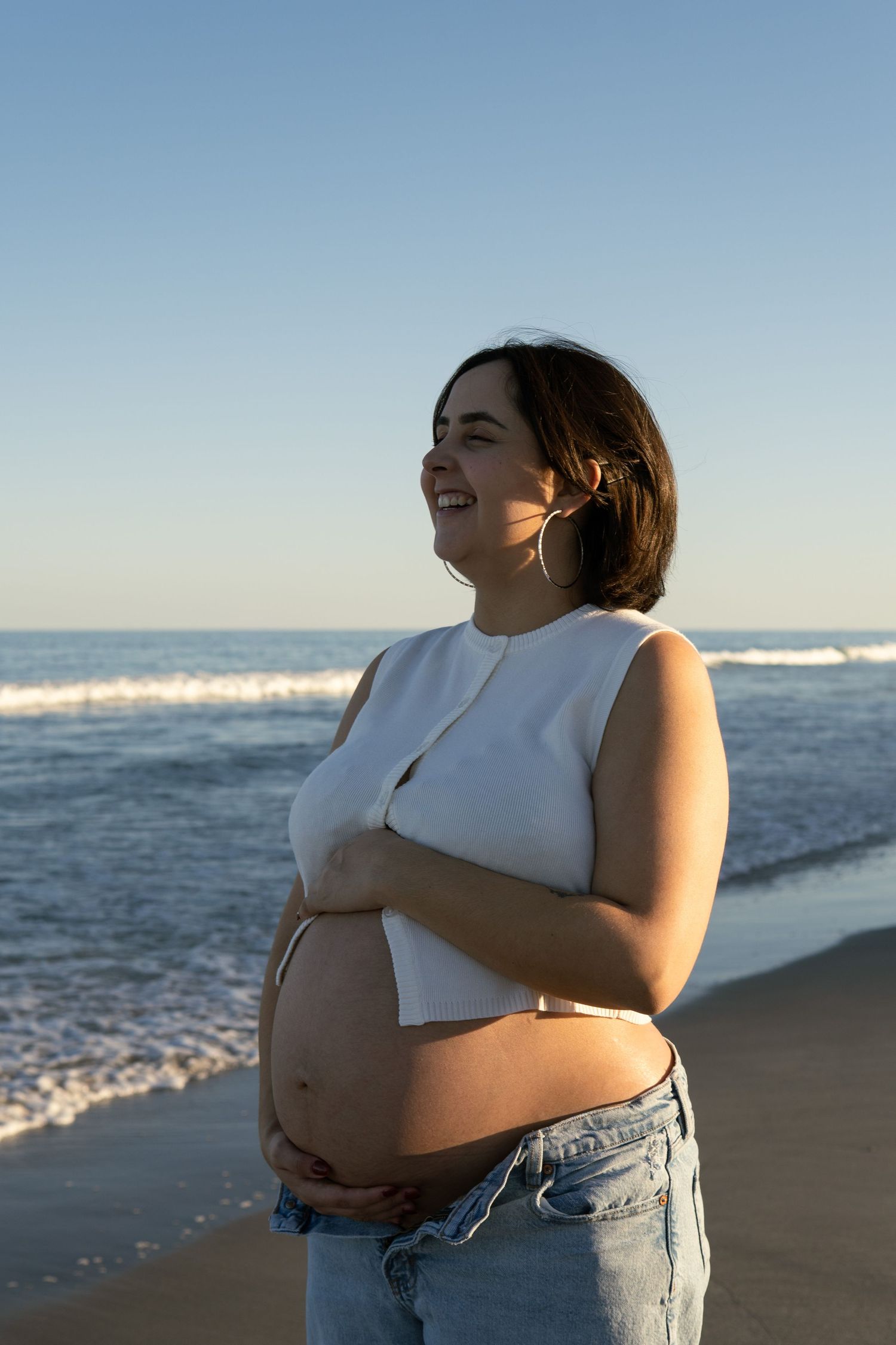 A smiling pregnant woman cradles her baby bump while standing on a sunny beach.