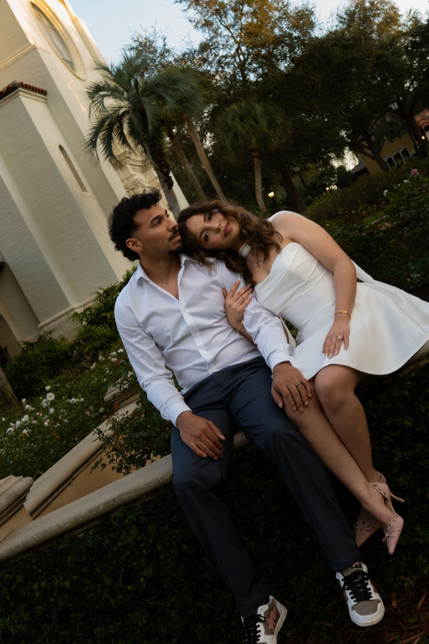 Couple sitting together on a ledge in a garden at sunset, woman in white mini dress sitting on mans lap smiling.