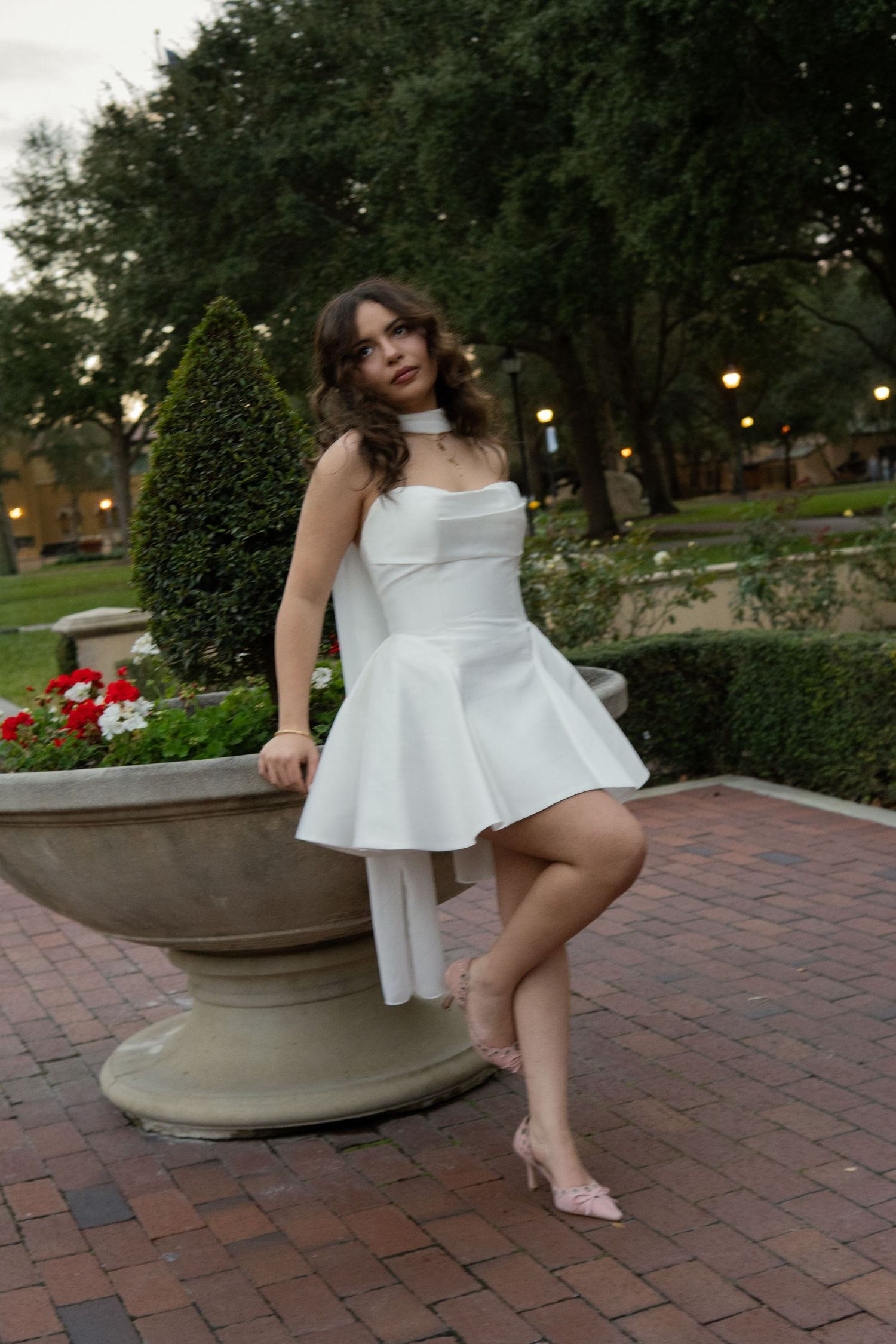 Woman in white structured mini dress posing beside a large stone planter in a garden at dusk.