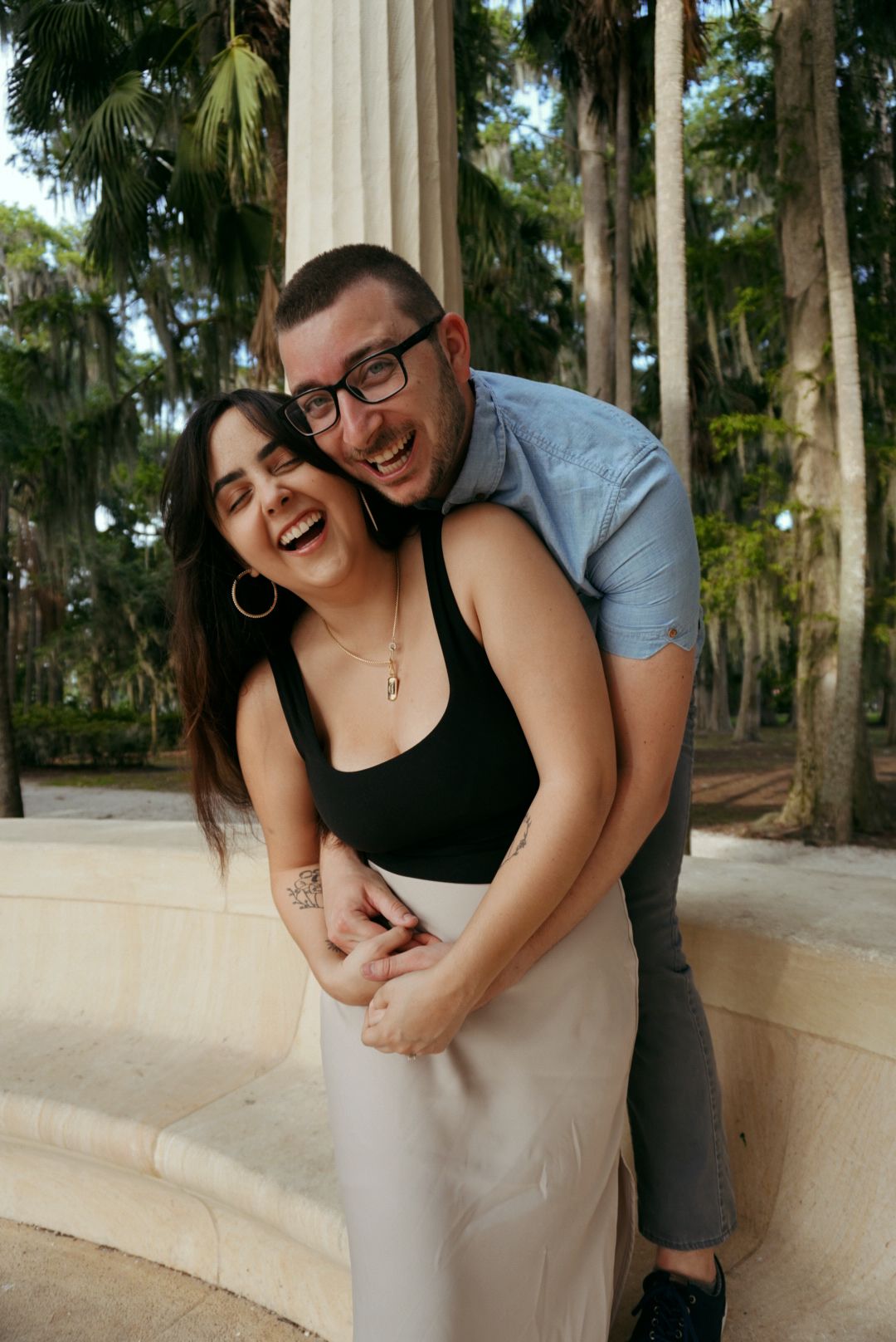 Joyful couple laughs together beside marble columns surrounded by palm trees in a park.