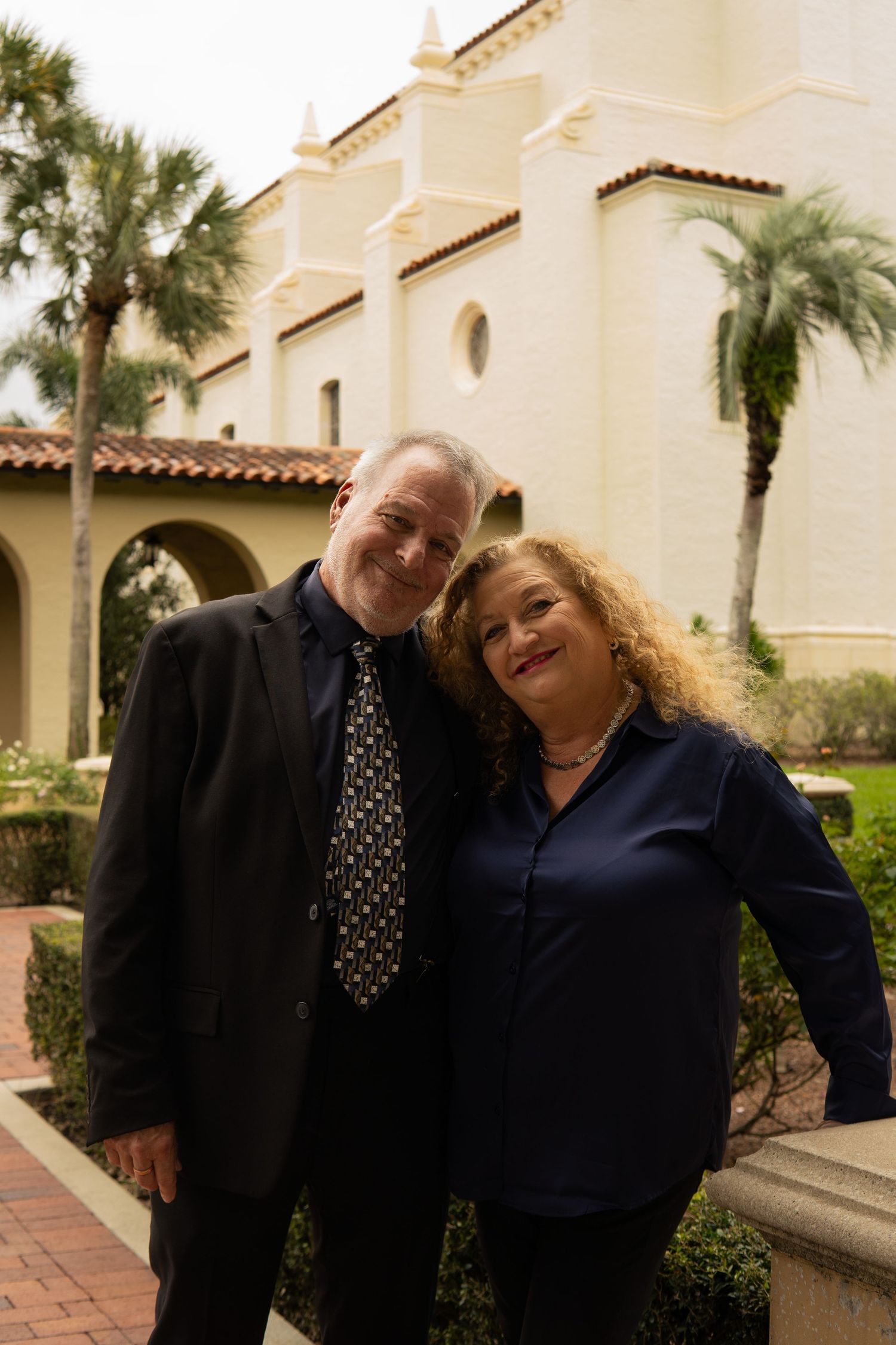 Smiling couple posing together outside a Spanish-style building with palm trees.