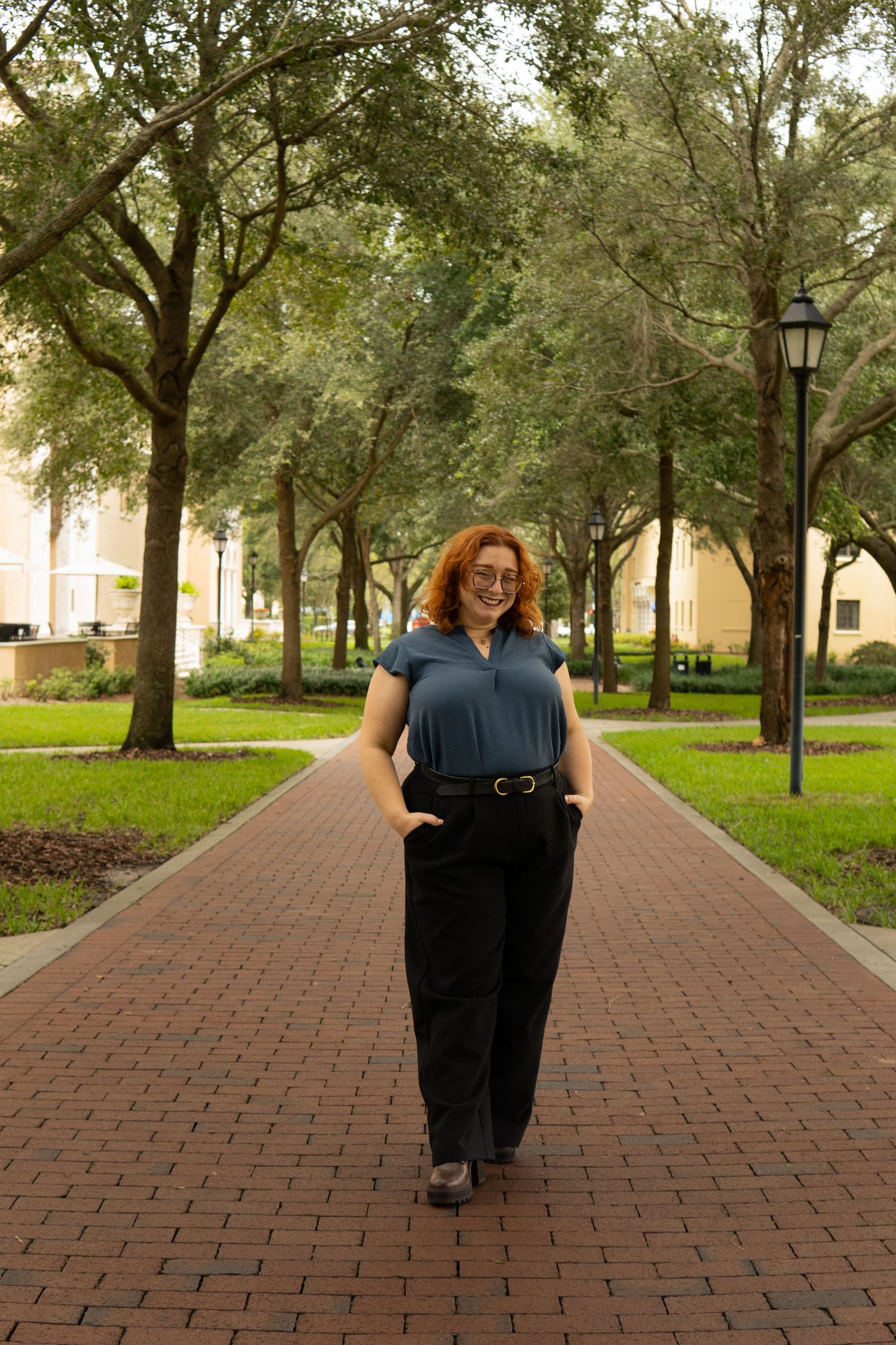 Red-haired woman in glasses walking confidently along a tree-lined brick pathway outdoors.