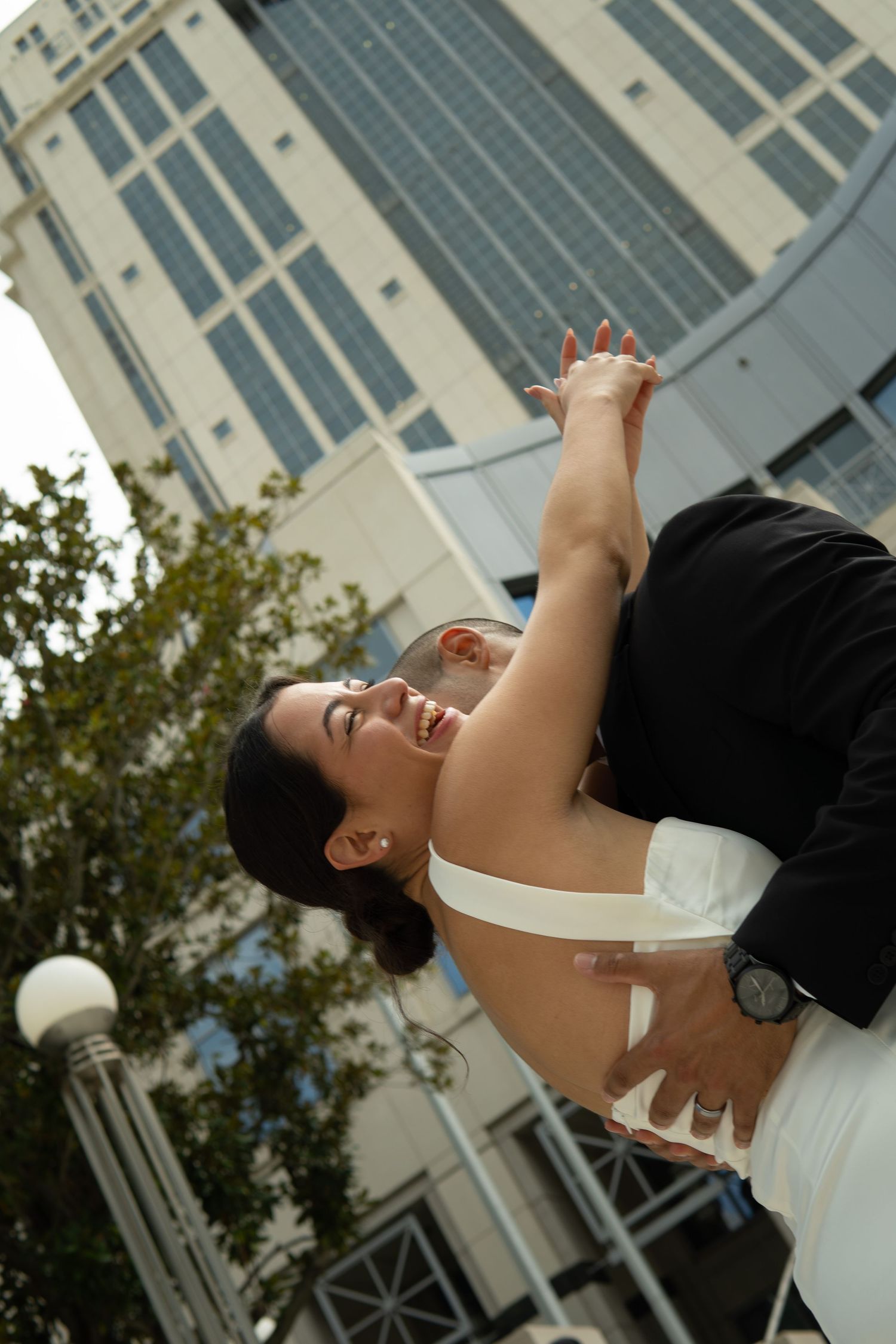 Groom joyfully lifting his laughing bride outdoors with a modern building in the background.