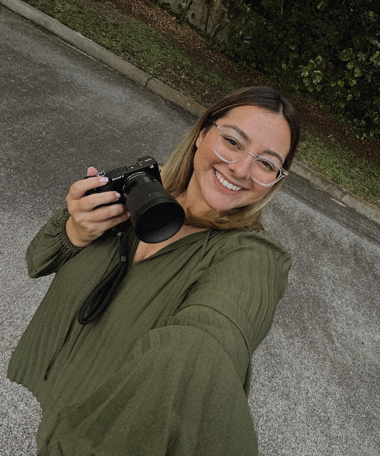 Smiling female photographer in green hoodie takes outdoor selfie while holding professional camera.