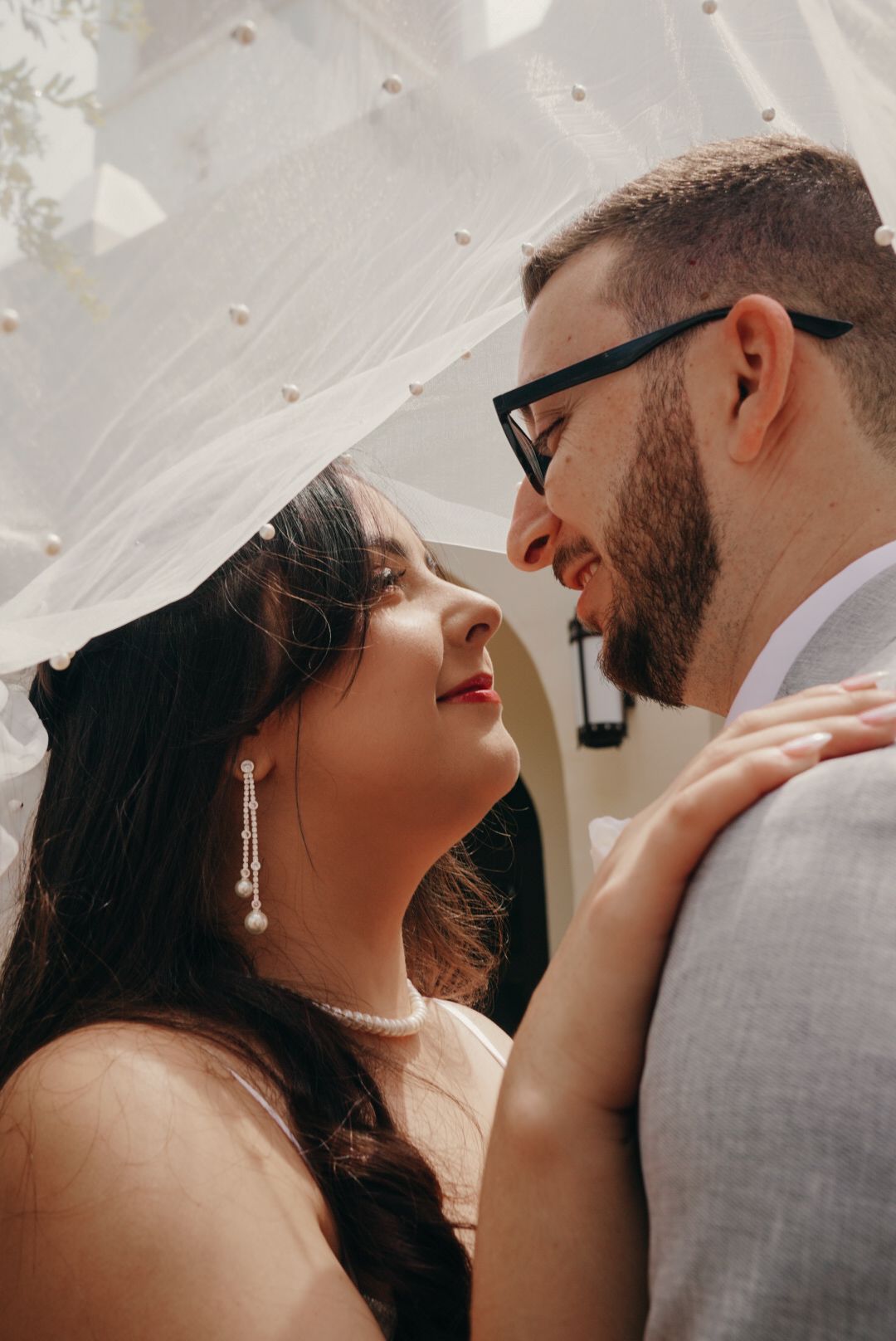Bride and groom sharing an intimate moment under a pearl-adorned veil, gazing into each others eyes.