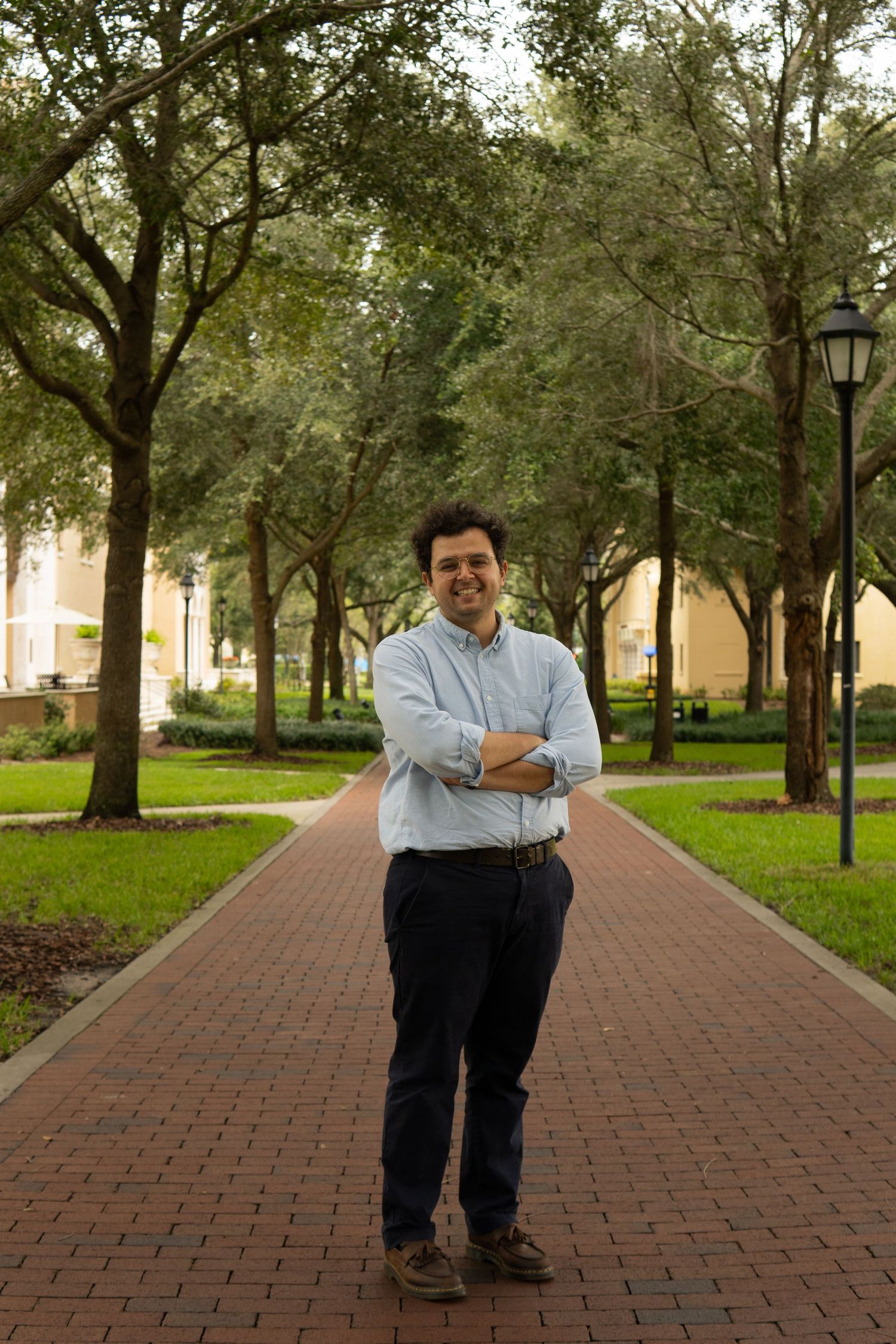 Cheerful man with arms crossed standing on a brick pathway surrounded by lush green trees.