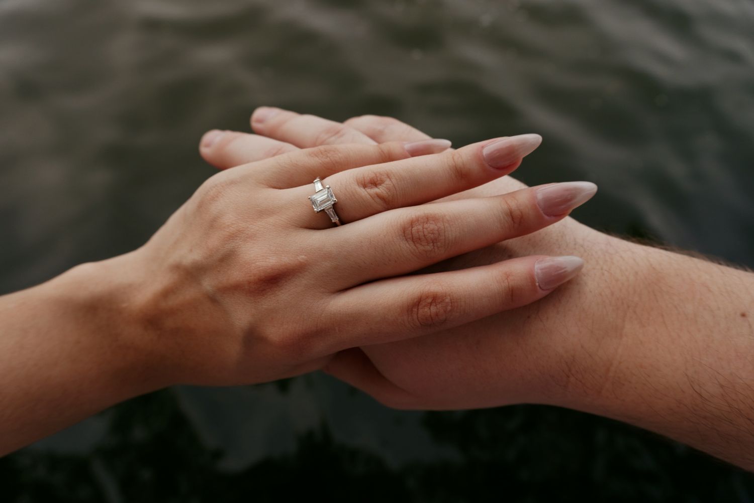 Close-up of an engagement ring on a woman's hand resting over her partner's hand by water.
