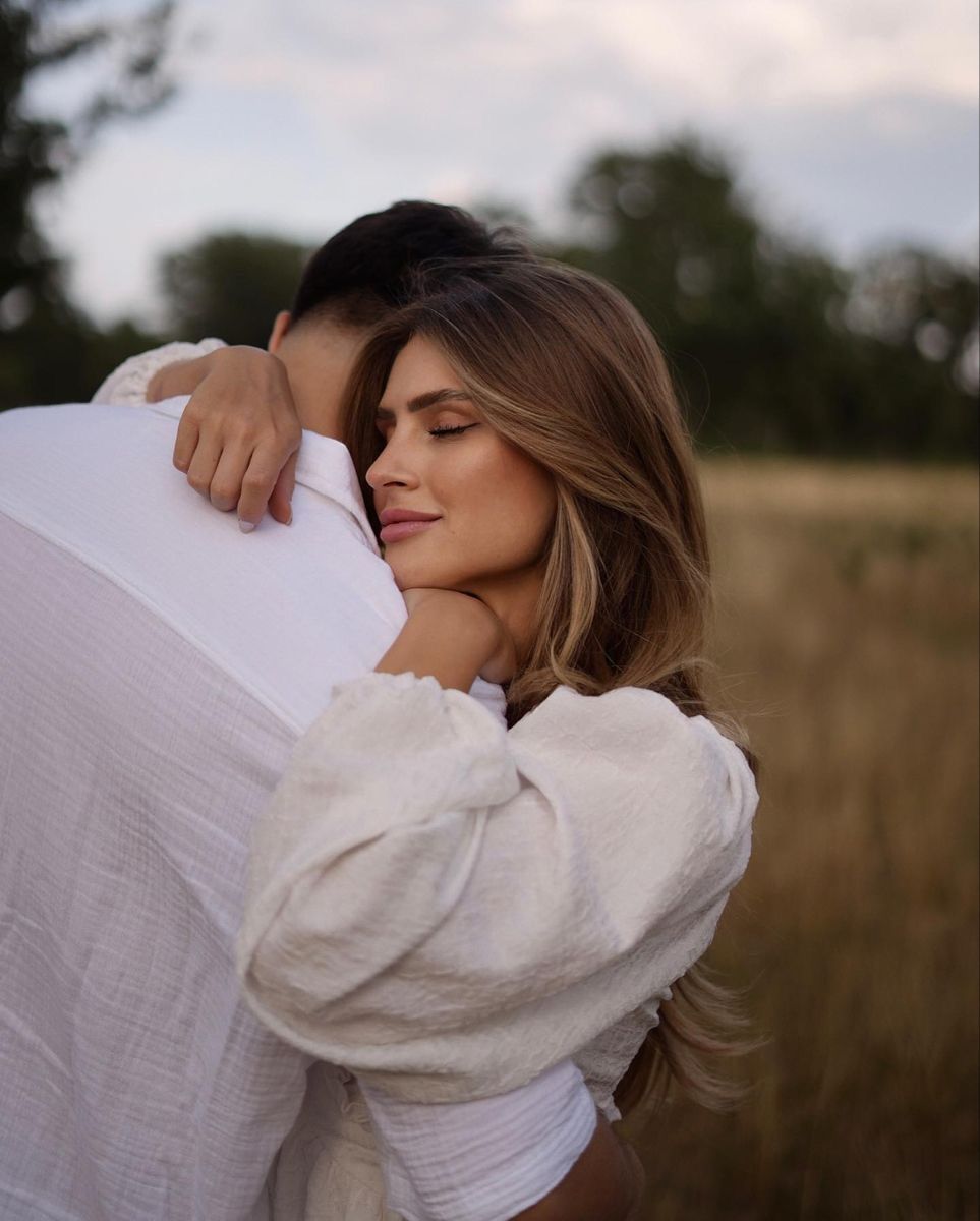 A romantic couple embraces in a sunlit field wearing white linen clothing during golden hour.