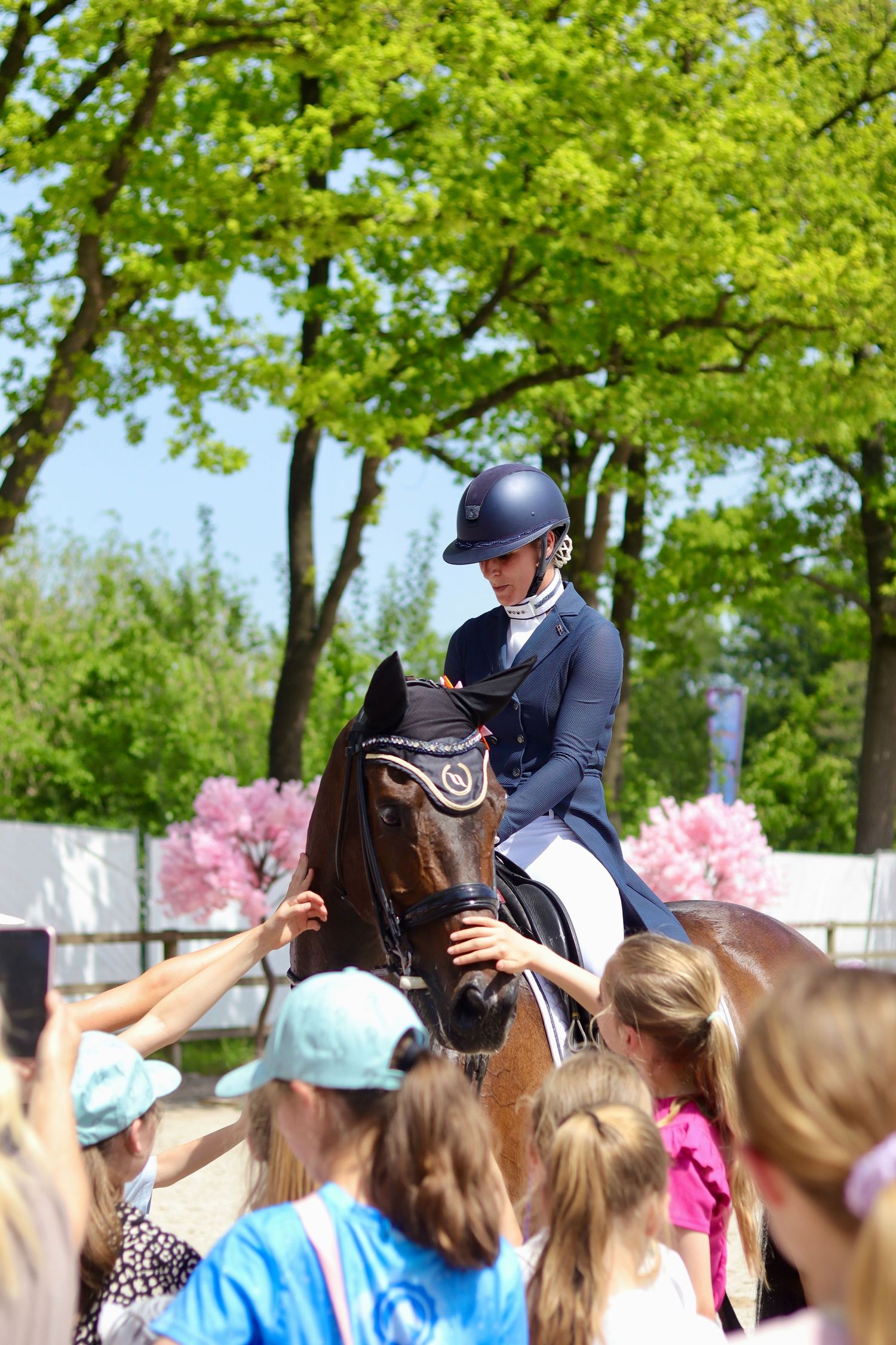 Equestrian rider in blue jacket and helmet on brown horse surrounded by children at outdoor competition.