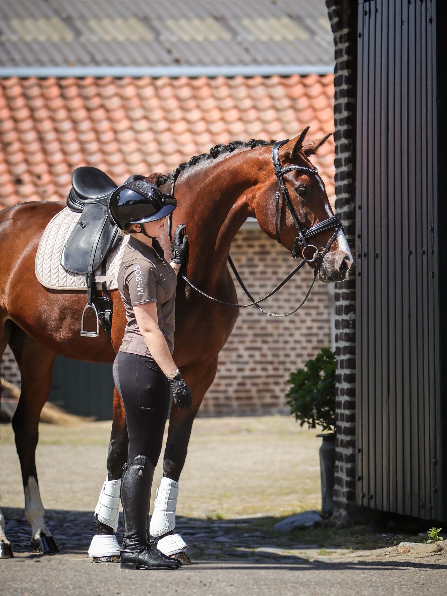 A woman in riding gear affectionately nuzzles her chestnut horse outside a stable.