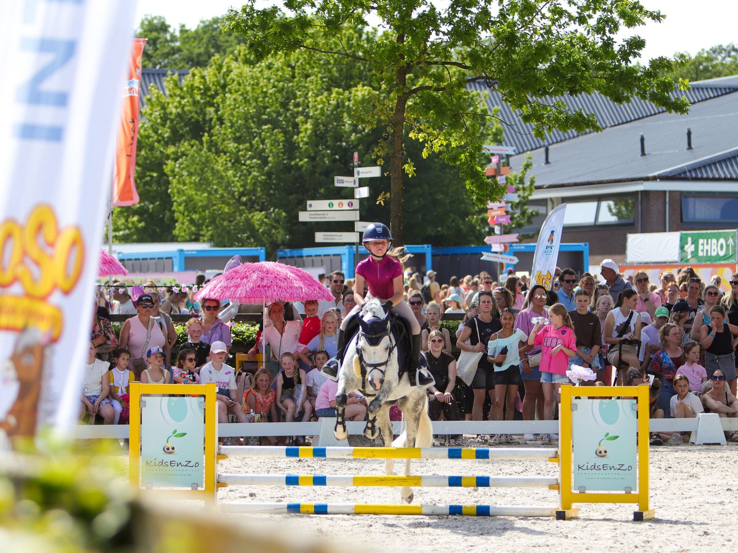 Rider on white horse jumping over colorful fence at outdoor equestrian show with large crowd watching on sunny day.