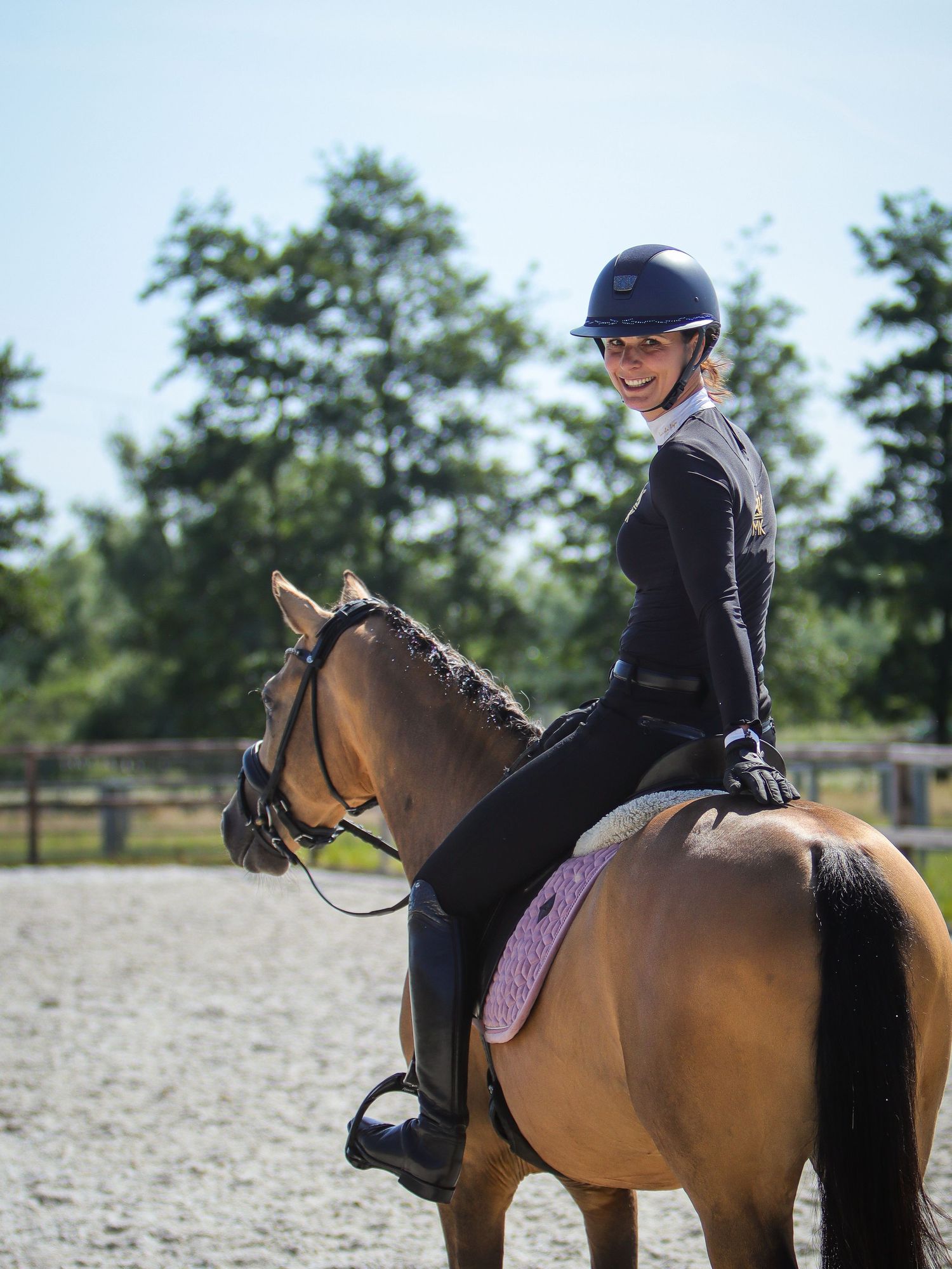 Smiling female equestrian in navy jacket and helmet riding a palomino horse in an outdoor arena.