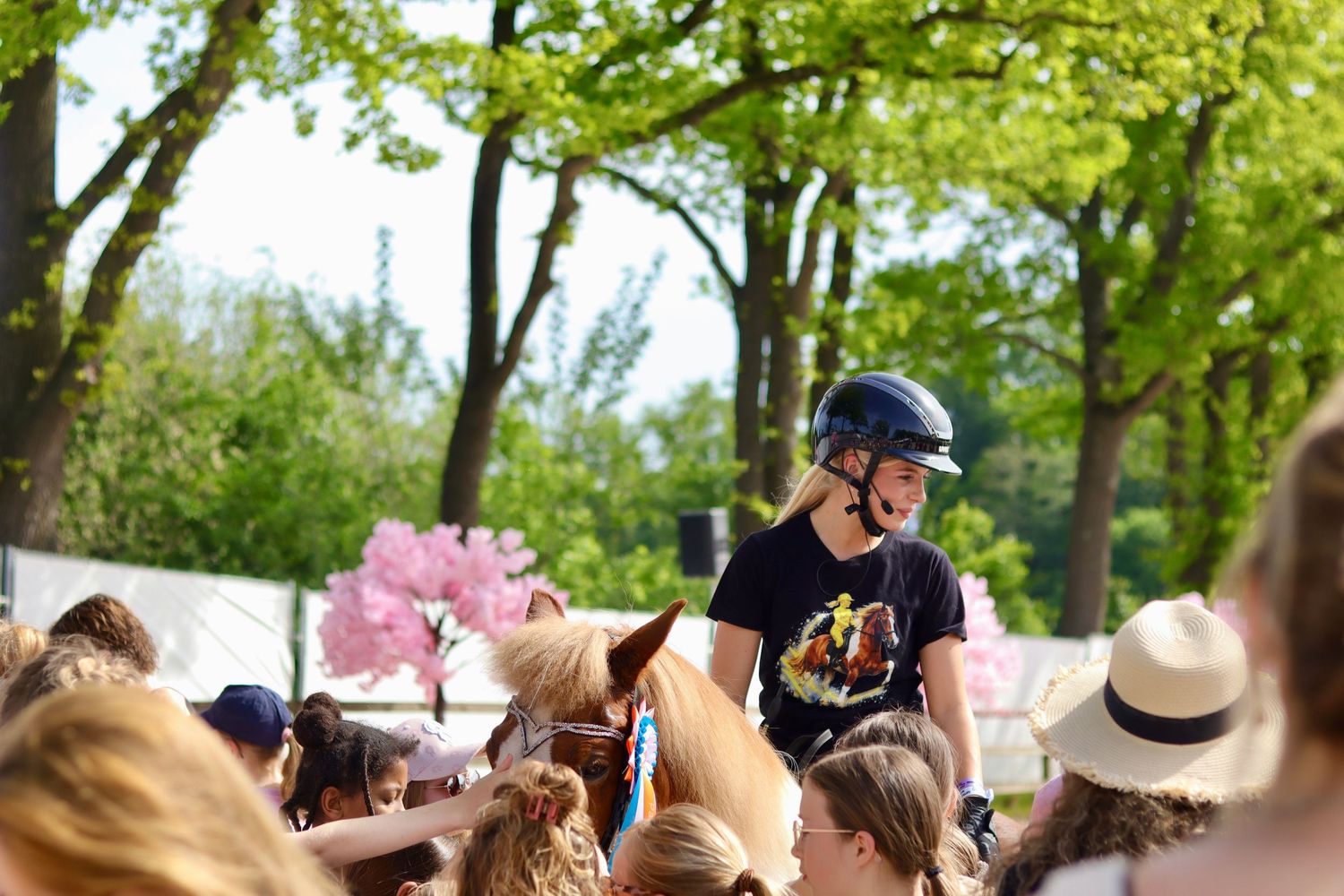 A rider in a black helmet on horseback surrounded by a crowd of children outdoors among green trees.