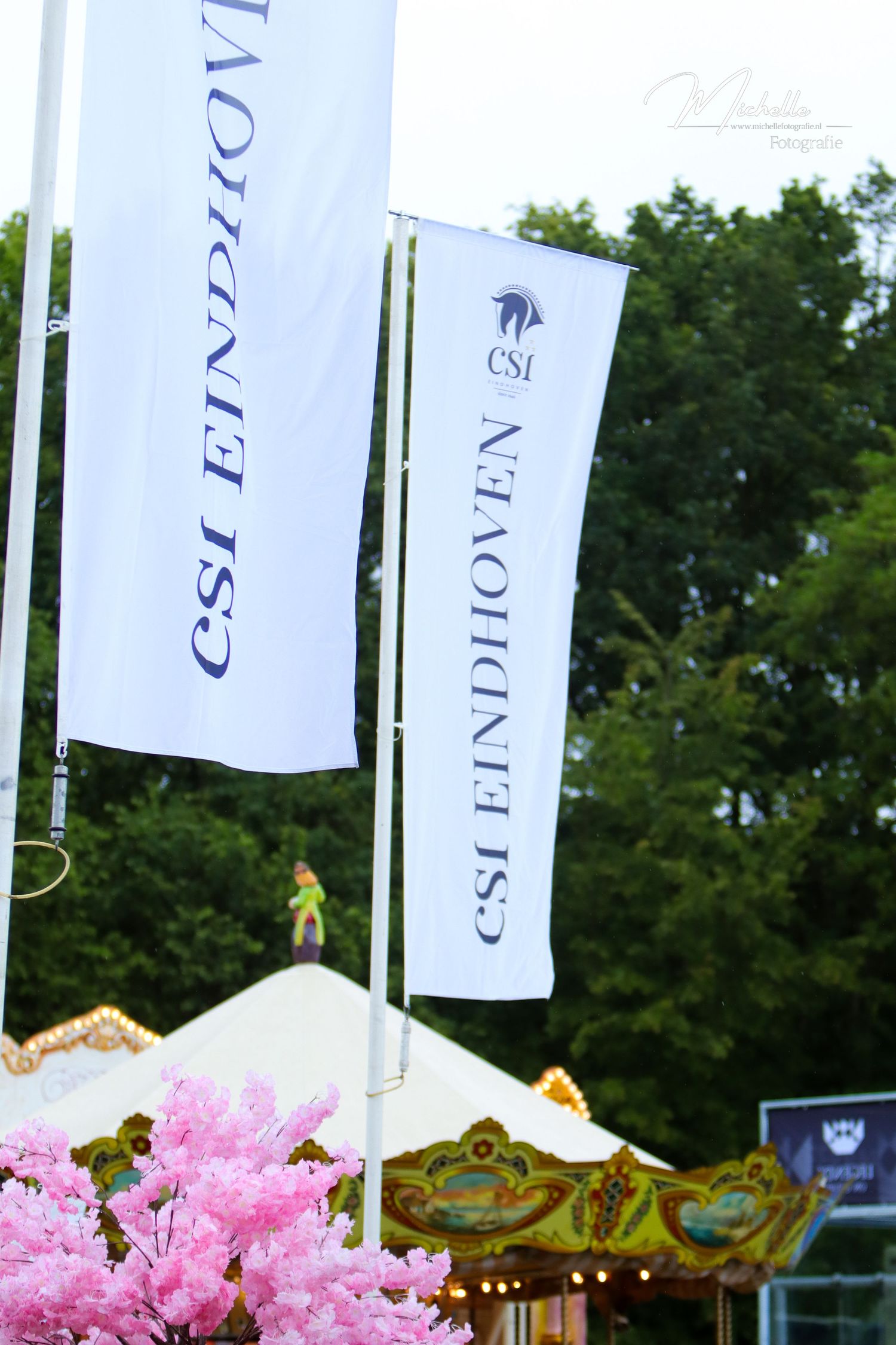 Two white CSI Eindhoven flags flying at an outdoor equestrian event with pink cherry blossoms and a carousel in background.