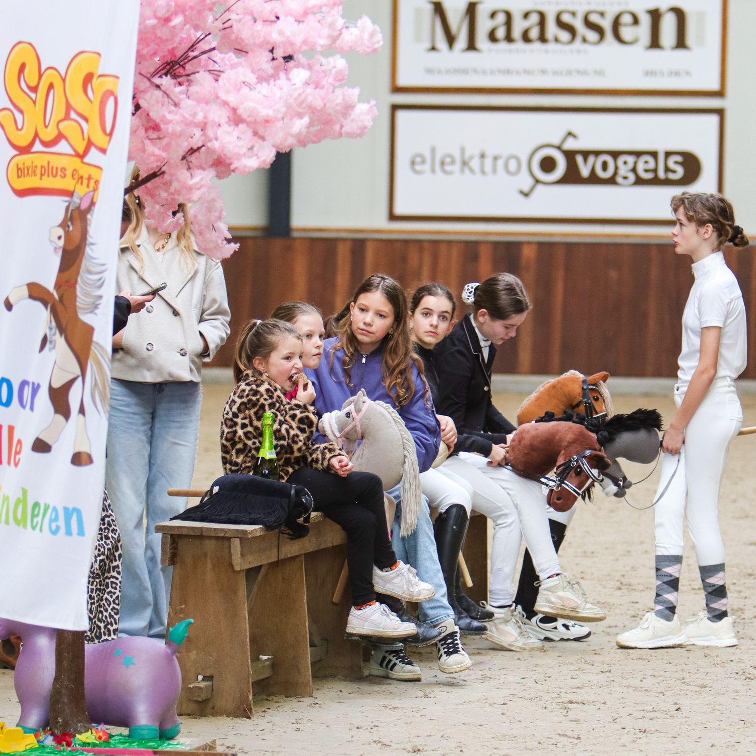 Children sitting on wooden benches at an indoor equestrian event with pink blossom decorations and sponsor signs.