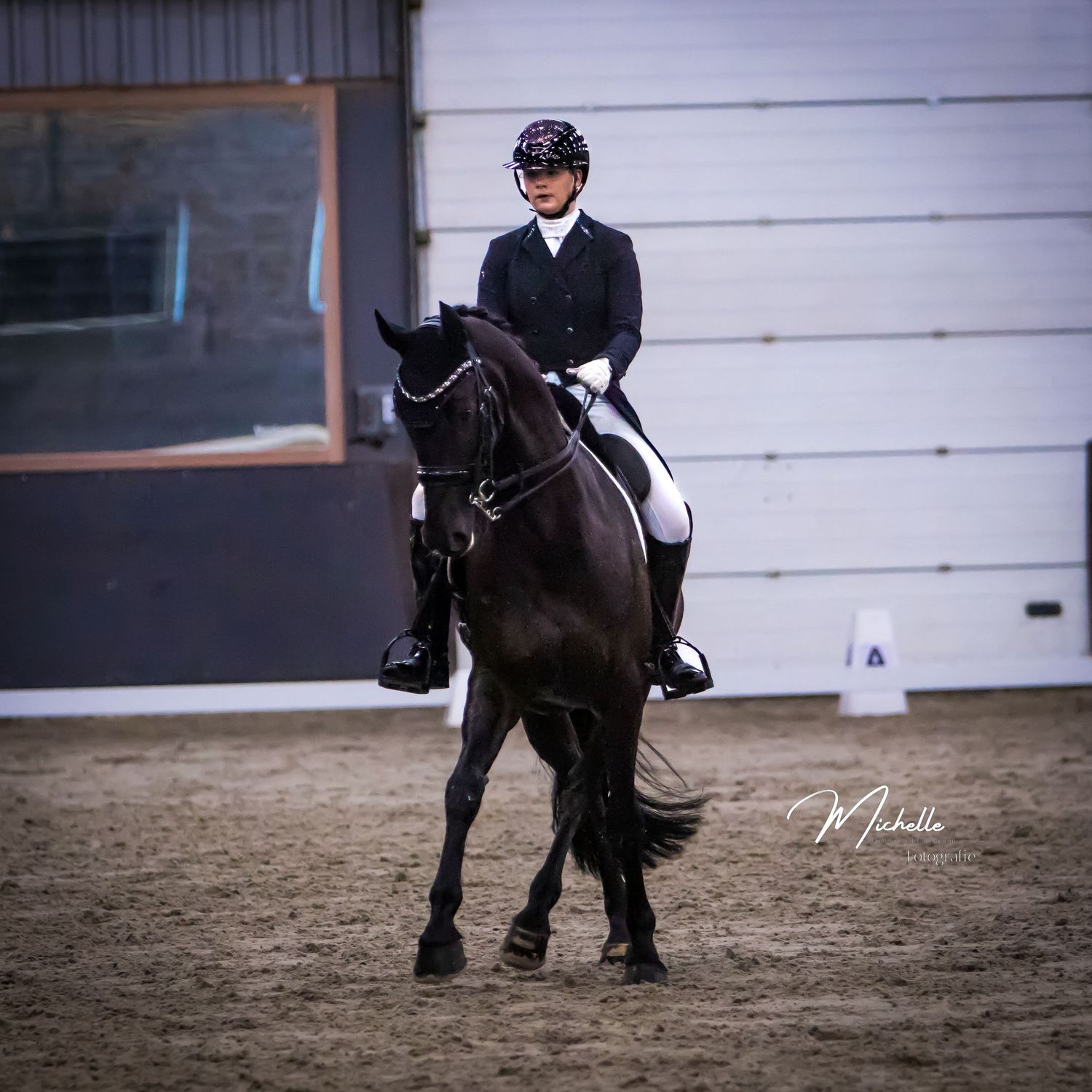 A rider in formal equestrian attire performs dressage movements on a black horse in an indoor arena.